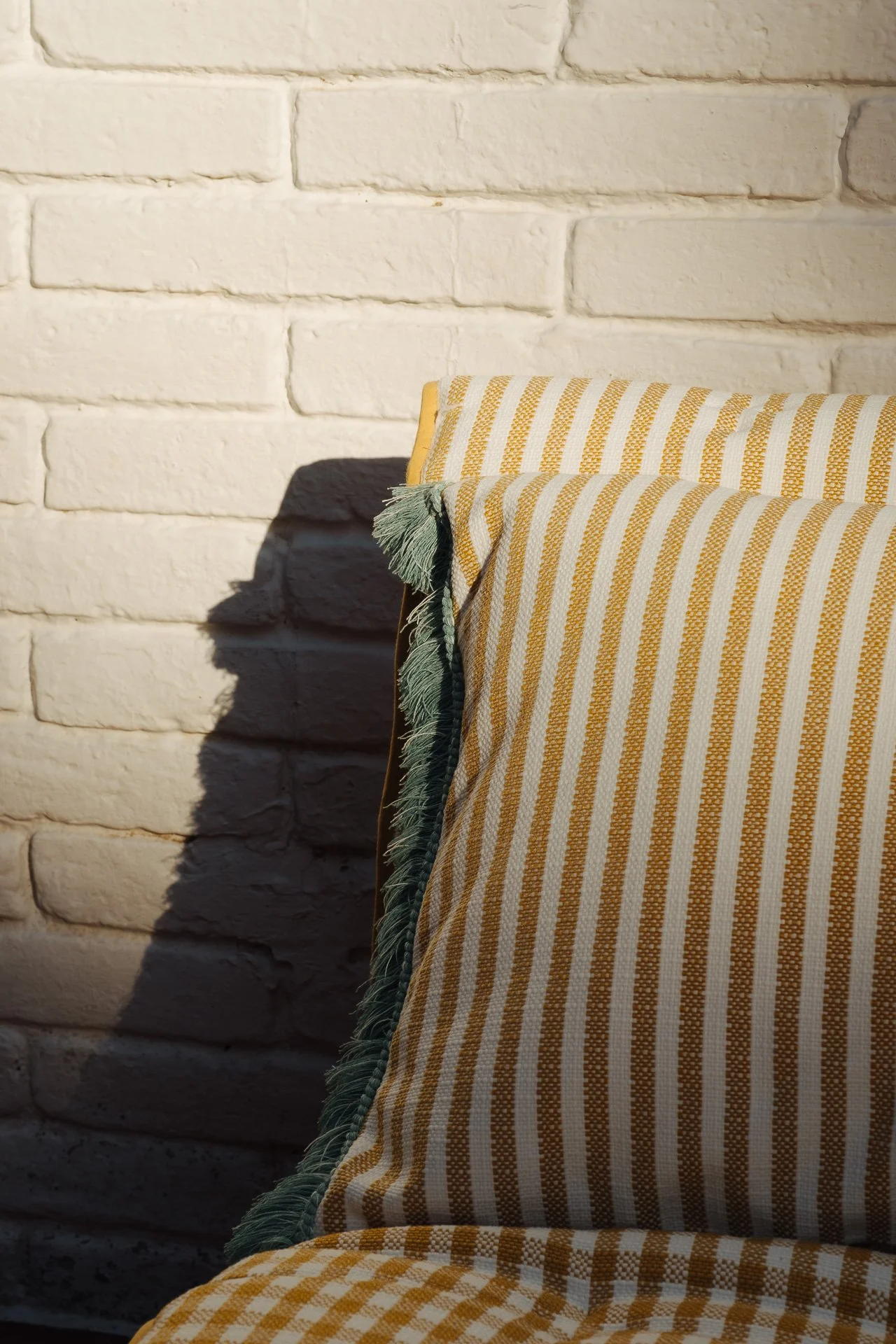 Fotografía comercial para Güell Lamadrid, Grupo Lamadrid, editores téxtiles, cojines con tela a rayas amarillas y blancas y un tapiz a rayas en tonos de amarillo y verde, en un sofá junto a una pared de ladrillos blanca con sombras en la luz solar.