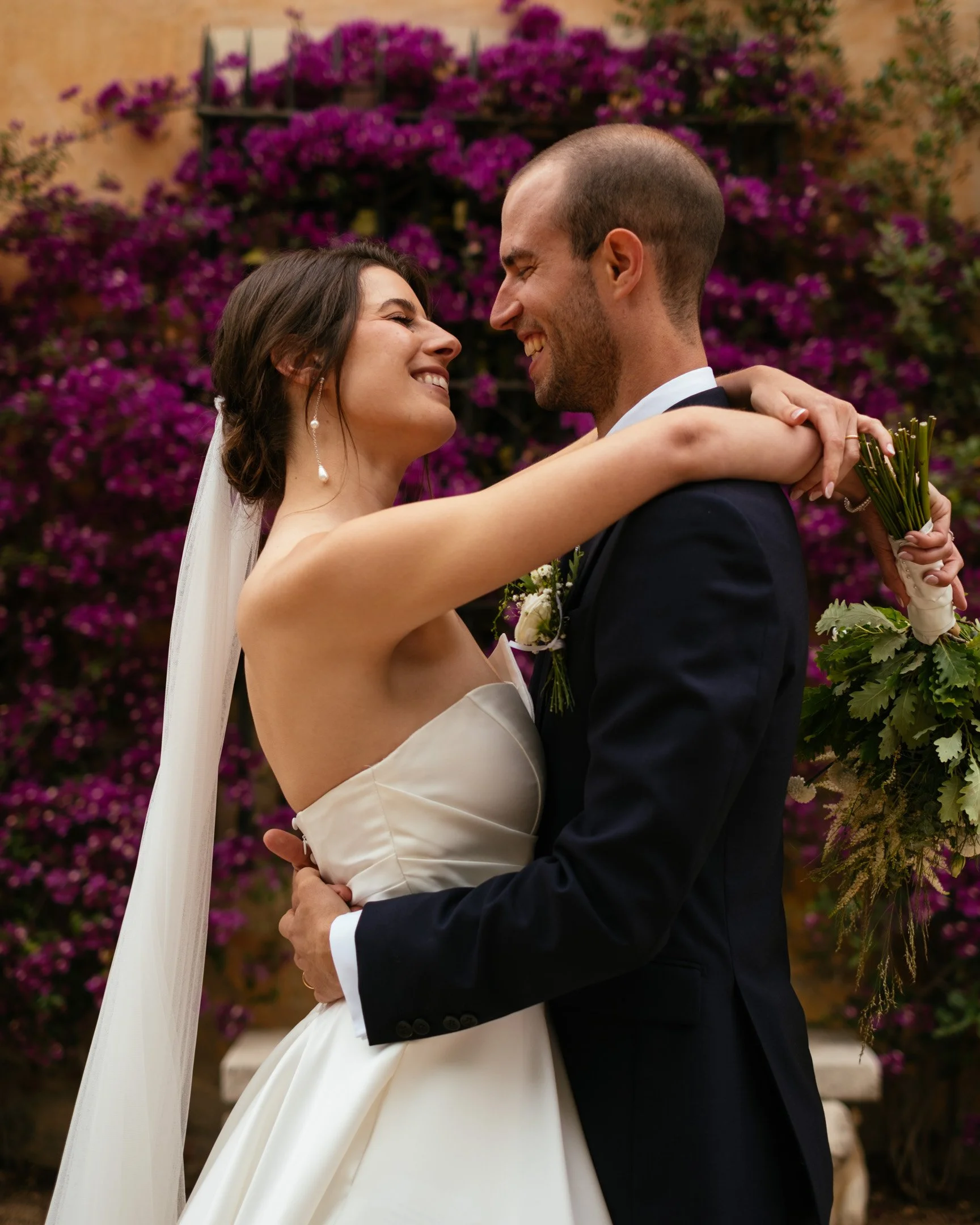 Pareja de boda sonriendo y abrazándose en medio de flores moradas.