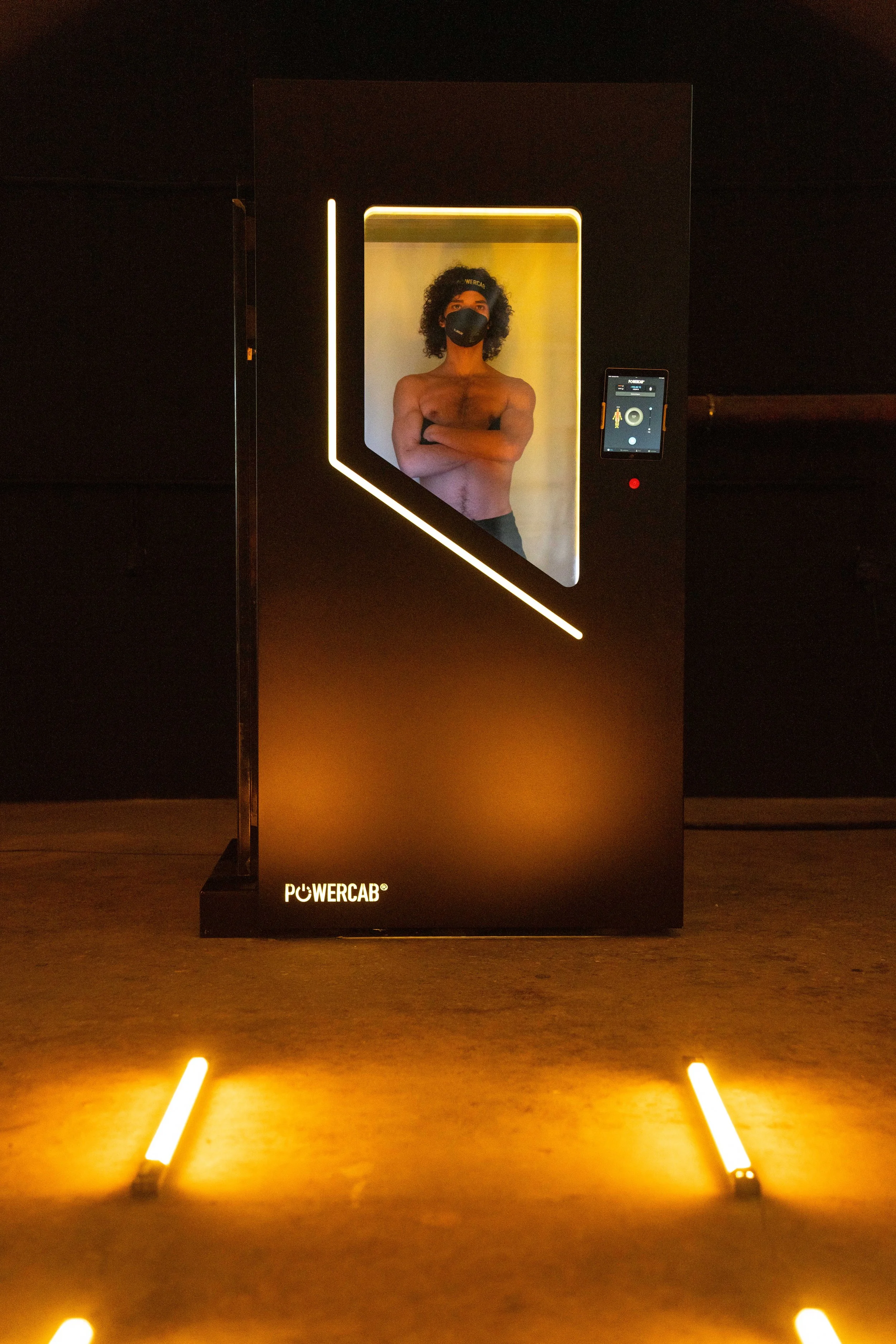 A person with curly hair, wearing a black face mask and headband, is inside a futuristic, illuminated photo booth labeled 'POWER CAB'. The person is shirtless, crossing their arms, and looking at the camera. Two yellow lights are on the floor in front of the booth.