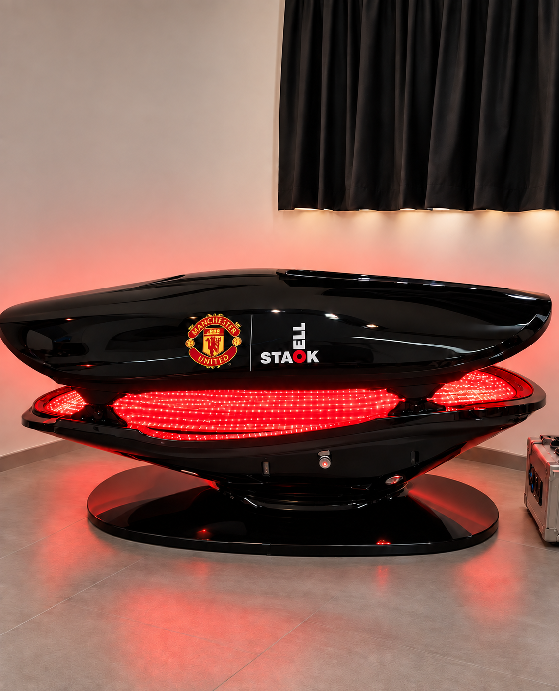 A futuristic medical pod with Manchester United and cell-stack logos, black glossy exterior, illuminated red interior, placed in a room with black curtains and beige walls.
