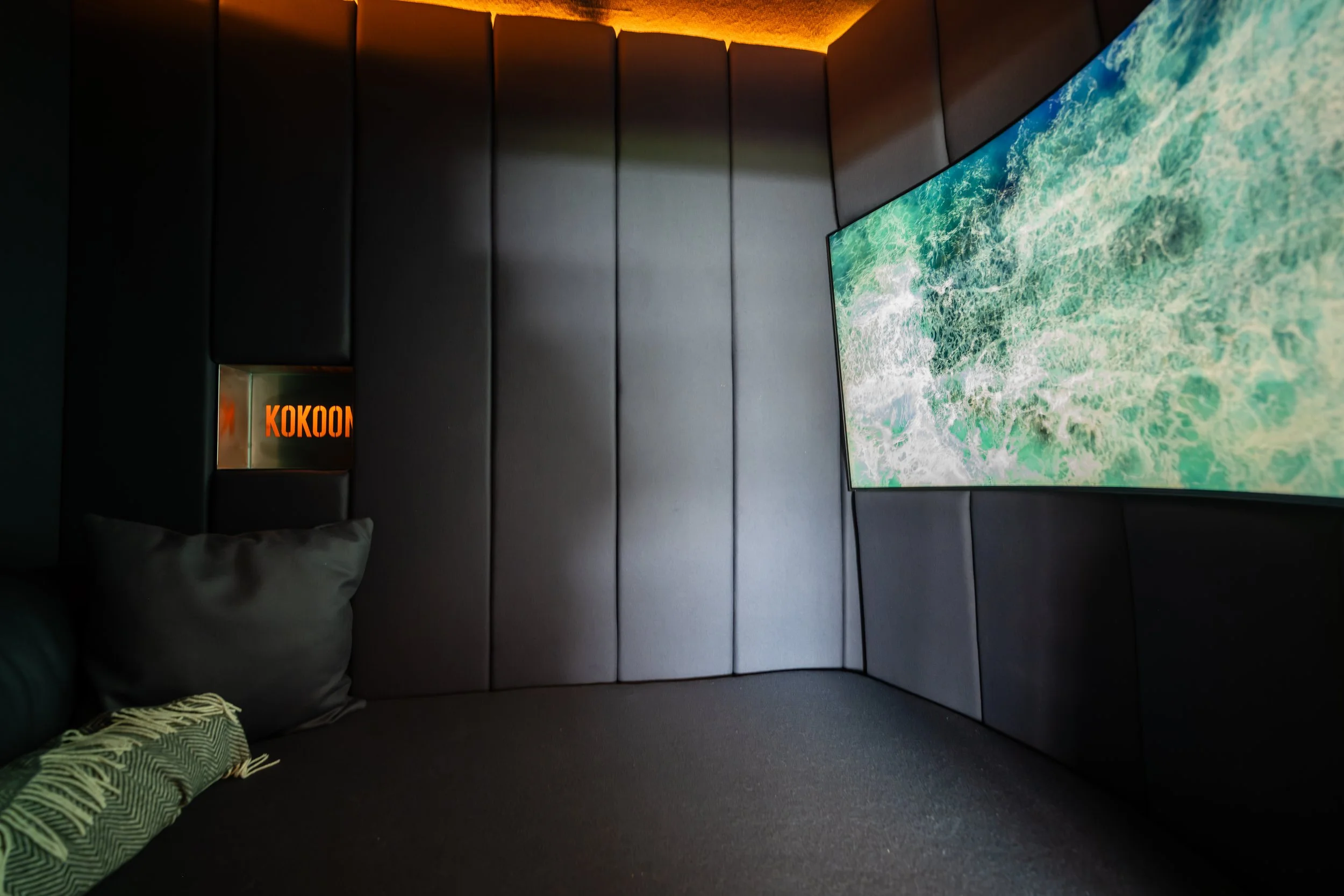 A modern dark-room with a large curved screen displaying a nature scene, black padded walls, a small illuminated sign that reads 'KOKOON,' a corner with pillows and a blanket. Kokoon unit by kloodos, at Manchester United