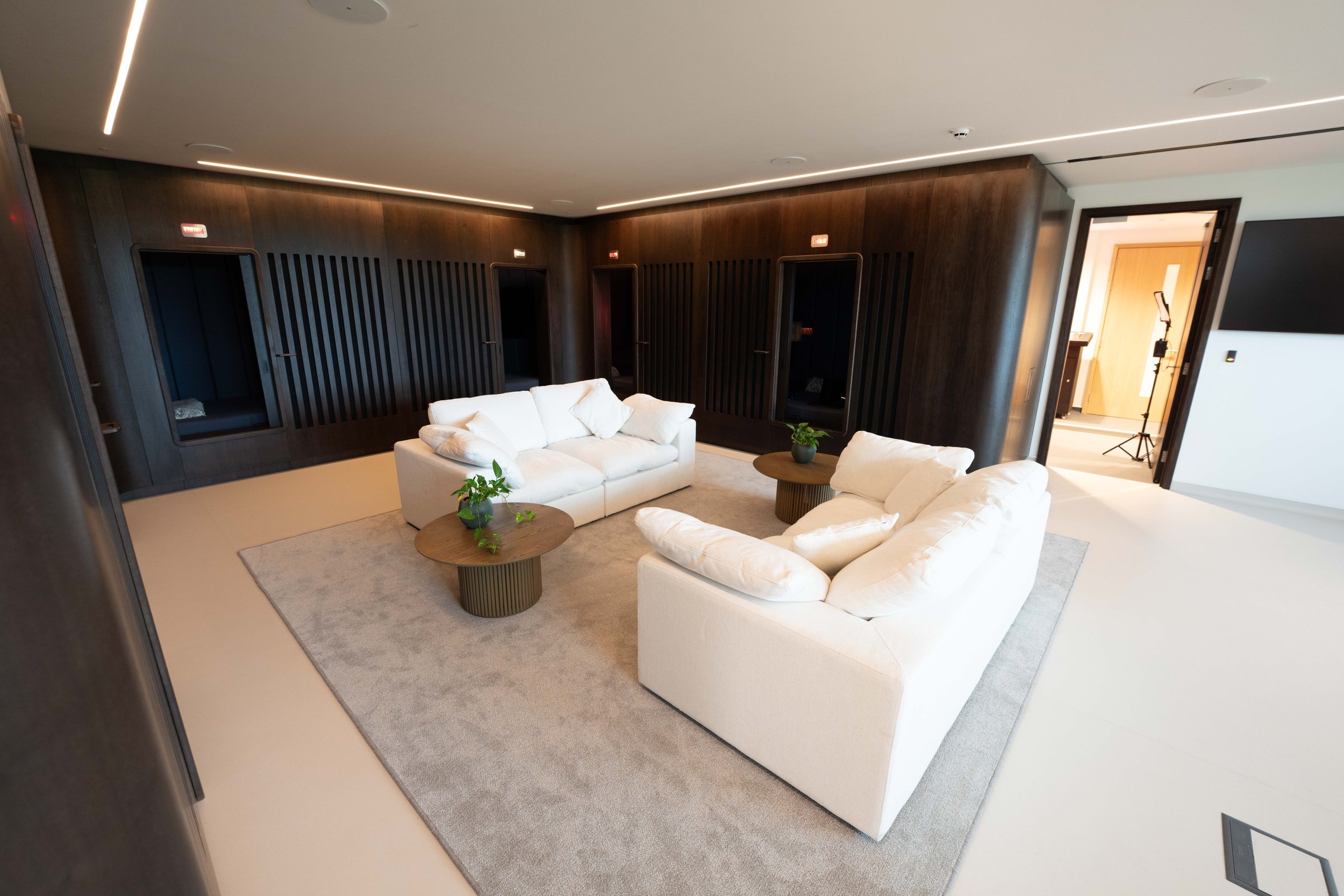 man united recovery room with a white sofa and two white armchairs arranged around a small coffee table with a plant. The room has dark wood-paneled walls with built-in niches, beige carpet, and linear ceiling lighting. kookon by kloodos