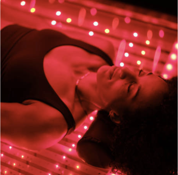 A woman with curly hair lying on her back, eyes closed, in a dark room illuminated by pink and red string lights.