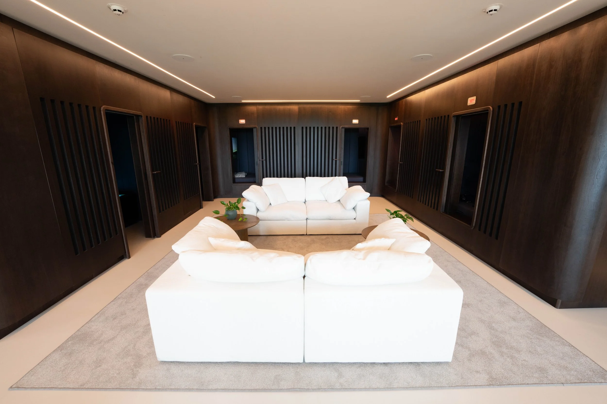 man united recovery room with a white sofa and two white armchairs arranged around a small coffee table with a plant. The room has dark wood-paneled walls with built-in niches, beige carpet, and linear ceiling lighting. kookon by kloodos
