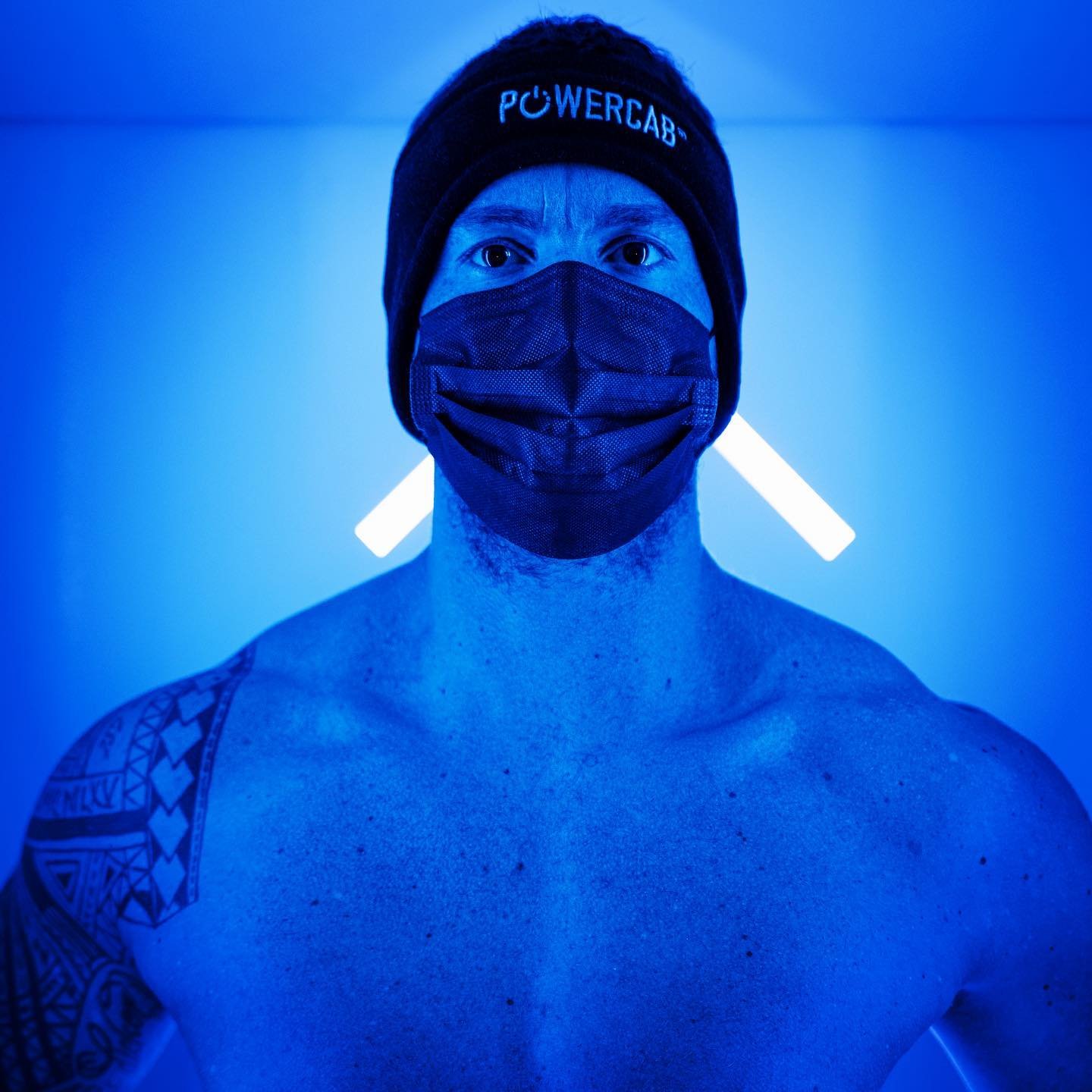 A shirtless person with tattoos on their arm and shoulder wearing a black beanie and a black face mask, illuminated by blue light, stood inside kloodos cryotherapy chamber