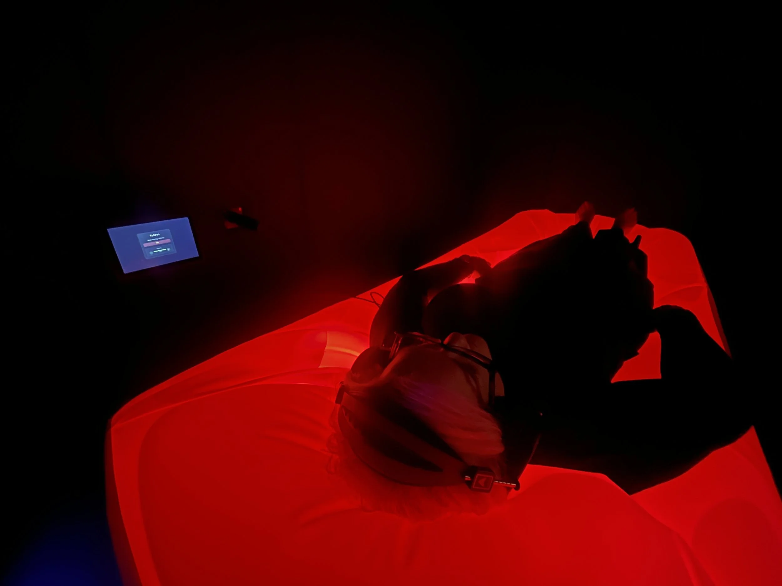 Person lying on k-float dry float  bed with red lights in a dark room, illuminated by a blue tablet screen showing a login interface to the kokoon-mini display.