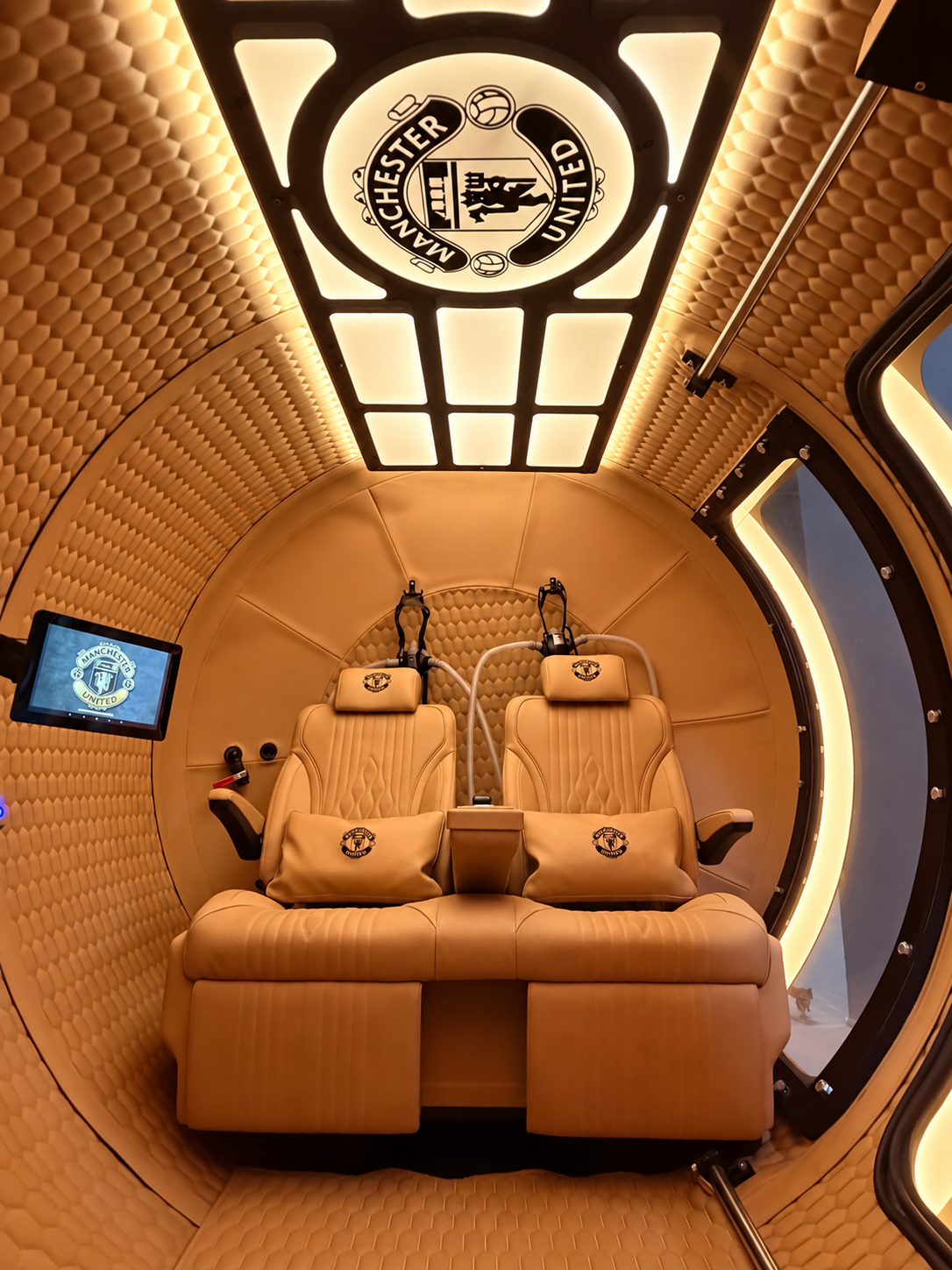oxy-stack hbot interior with Manchester United branding, featuring beige leather seats, quilted wall panels, and illuminated Manchester United logos on the ceiling and a small screen.