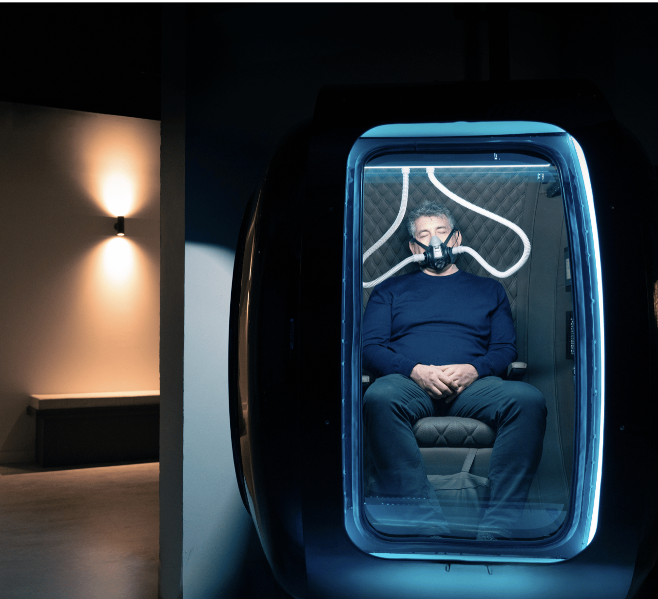 A man sitting inside an oxy-stack hbot chamber with a breathing mask, illuminated by blue LED lights.