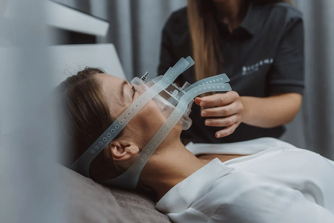 A woman lying on a bed wearing a mask and IHHT gear with tubes, while a healthcare worker prepares her.