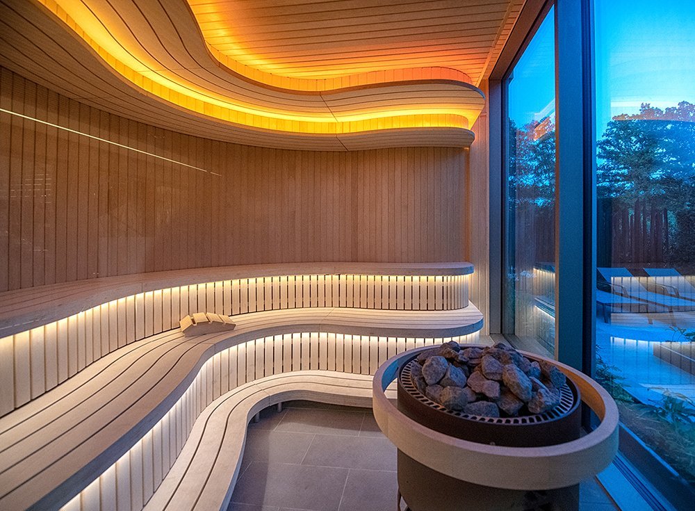 Modern sauna room with curved wooden benches illuminated by warm strip lighting, a large window showing outside trees, and a heater with rocks.