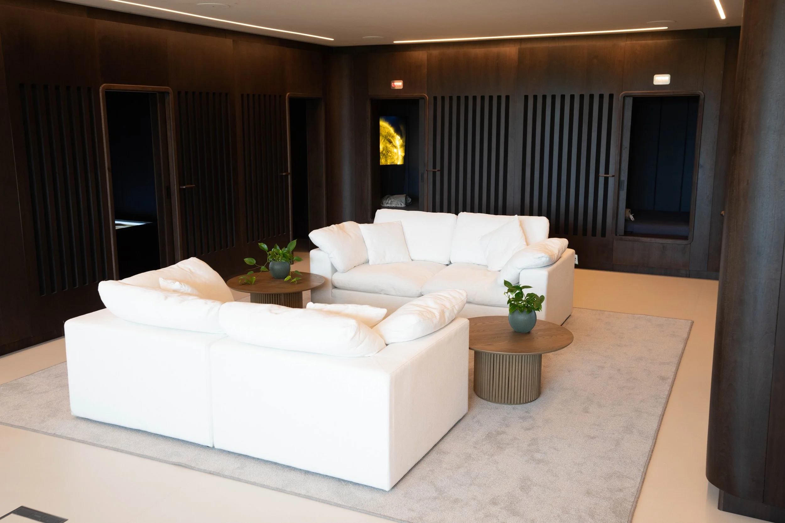 man united recovery room with a white sofa and two white armchairs arranged around a small coffee table with a plant. The room has dark wood-paneled walls with built-in niches, beige carpet, and linear ceiling lighting. kookon by kloodos