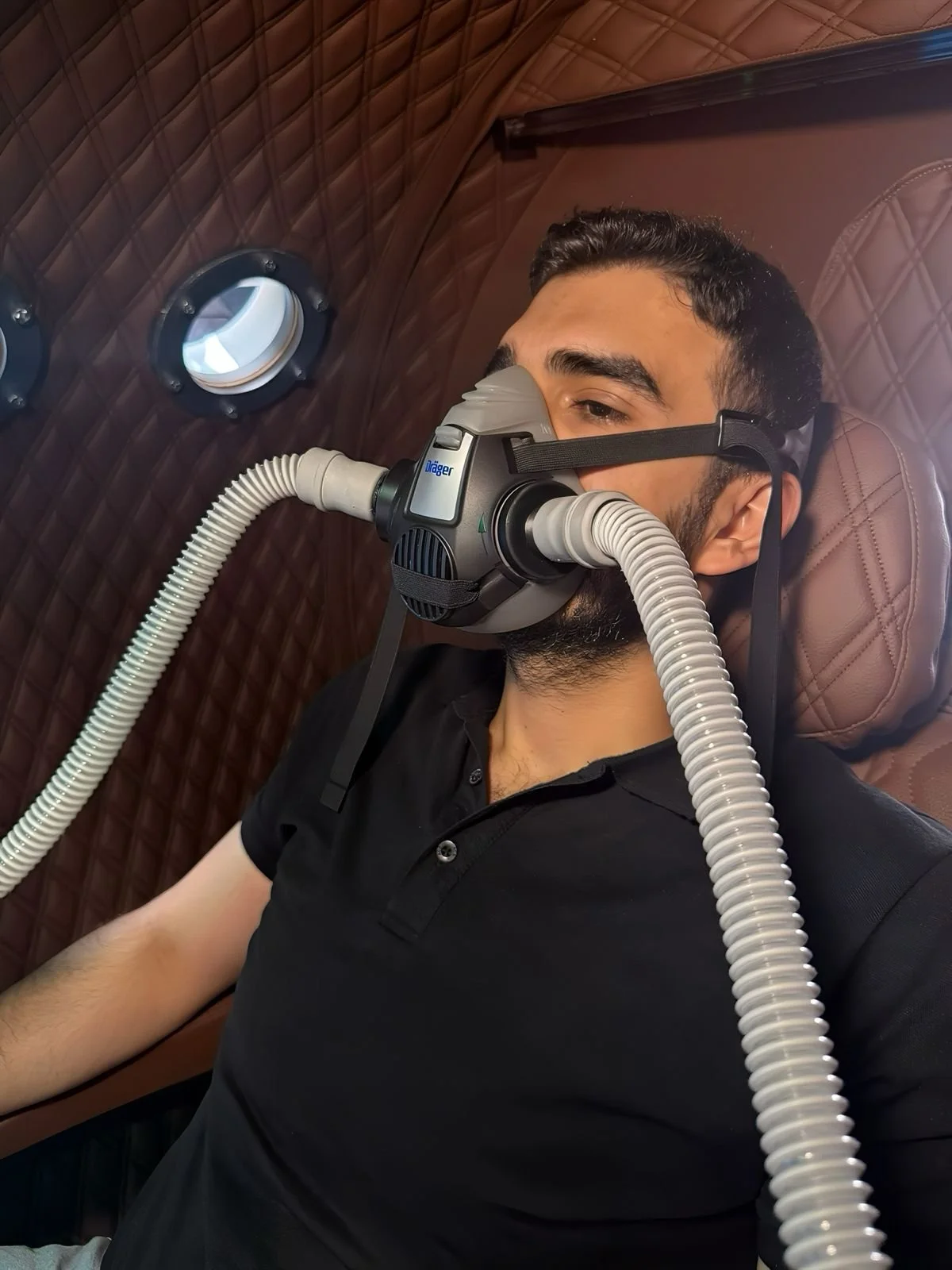 A man lying on a padded, quilted surface, wearing a mask with tubes connected, hbot therapy inside oxy-stack