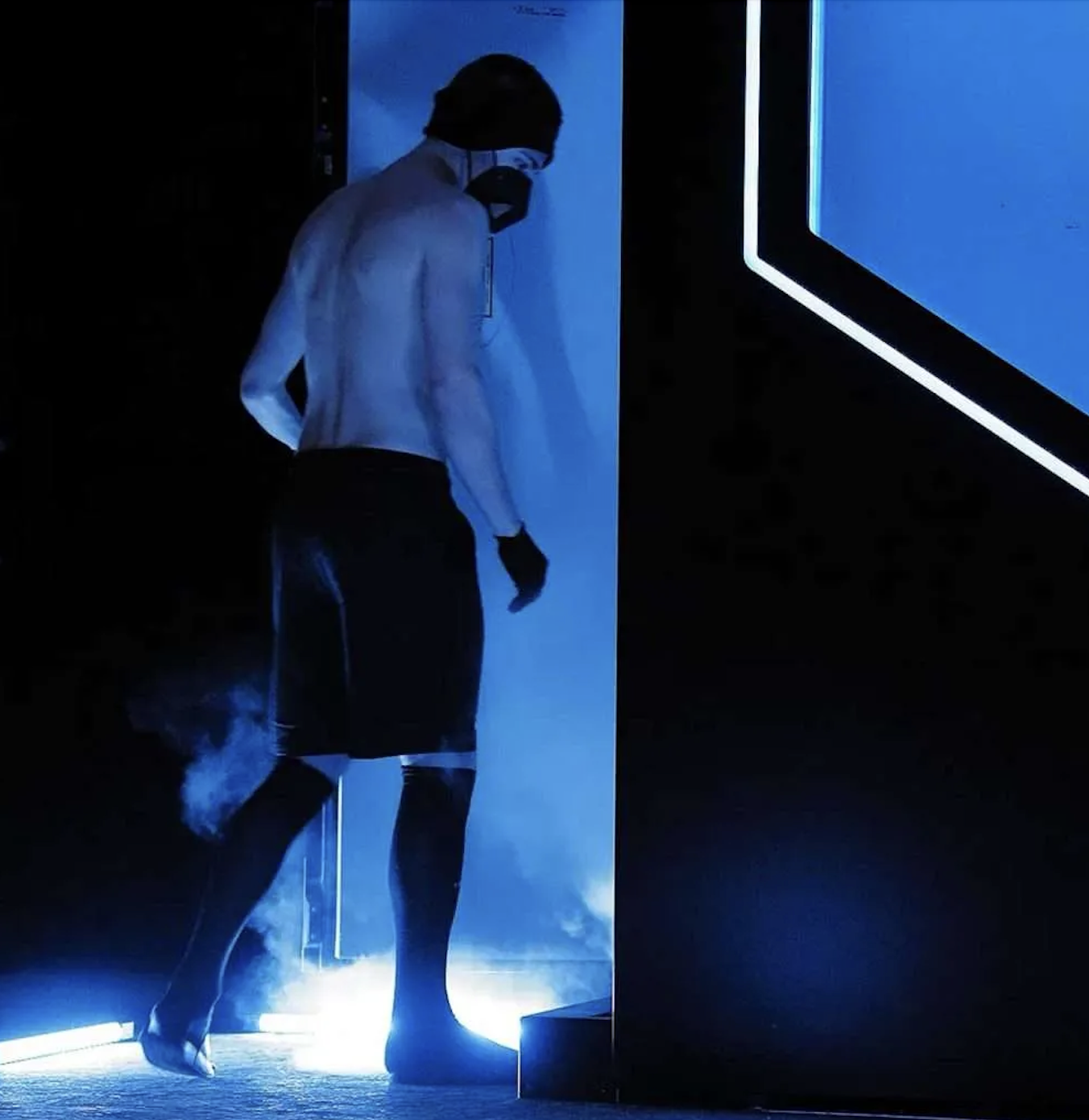 Person dressed in black and white clothing walking into a cryotherapy chamber illuminated by blue light.