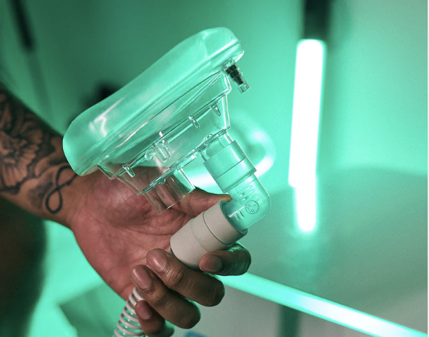 A person holding a small ihht device with clear plastic parts and a gray-colored connector pipe, against a background with green lighting.