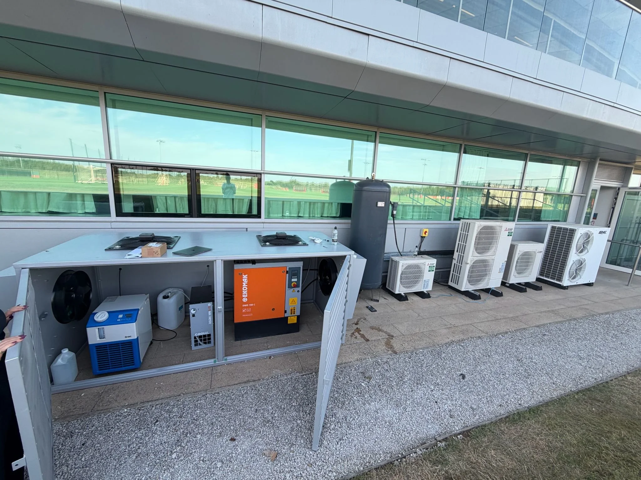 Outdoor view of a building with large windows, showing a green field and sports fields in the background. There is a table with various electronic and industrial equipment, including air conditioning units, a water bottle, and boxes. The scene appears to be on a balcony or terrace with a gravel floor.