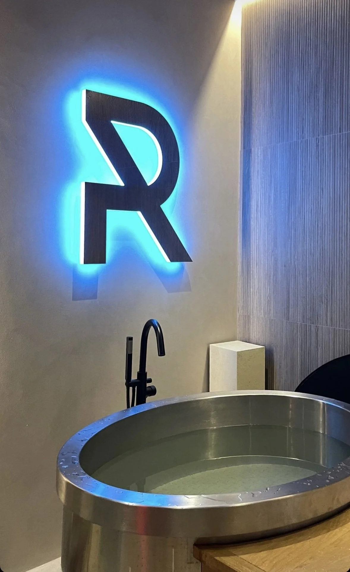 A stainless steel tub filled with water, with a modern black faucet above it. In the background, a large illuminated sign of the letter R with blue backlighting is mounted on the wall. The wall has a mix of plain and textured surfaces.
