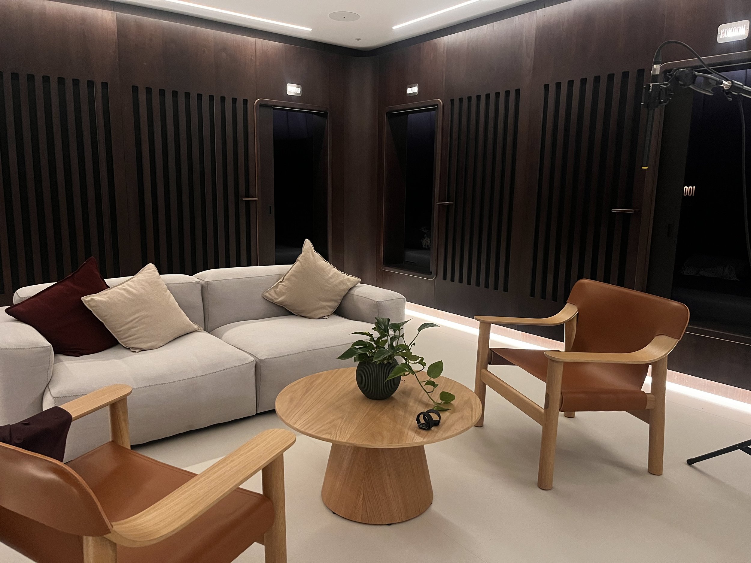 A modern lounge with a white sofa decorated with beige and red pillows, a wooden coffee table with a potted plant, and two contemporary wooden chairs, against a dark wood-paneled wall with doorways. Kokoon by kloodos 