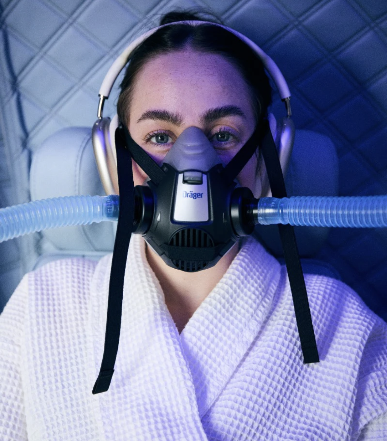 Person in a white robe wearing a breathing mask with tubes and headphones, lying on a cushioned surface.