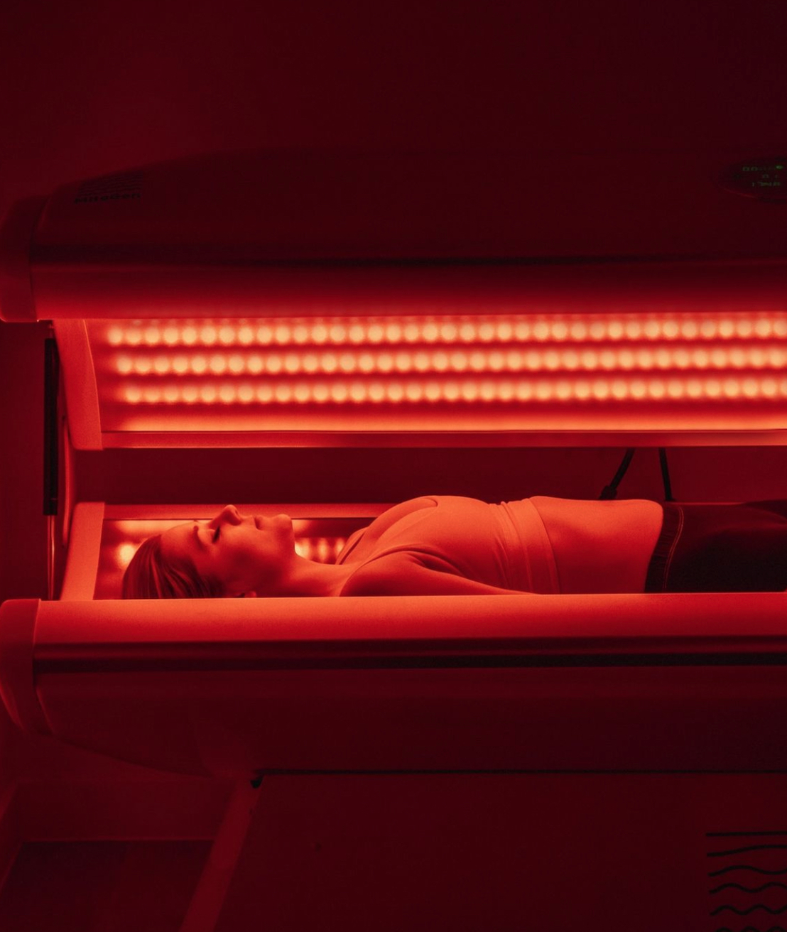 A woman lying inside a red cell-stack gen 2 pbm bed with bright red lights illuminating her face and body.