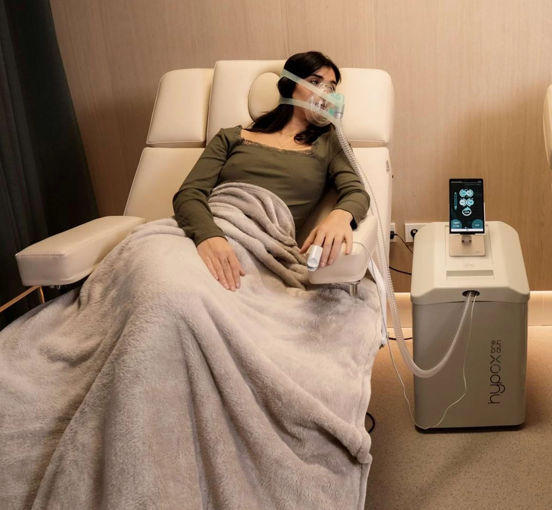 Woman lying on a medical chair wearing a breathing mask connected to a machine, with a remote control in hand, covered with a beige blanket, in a room with wooden walls.