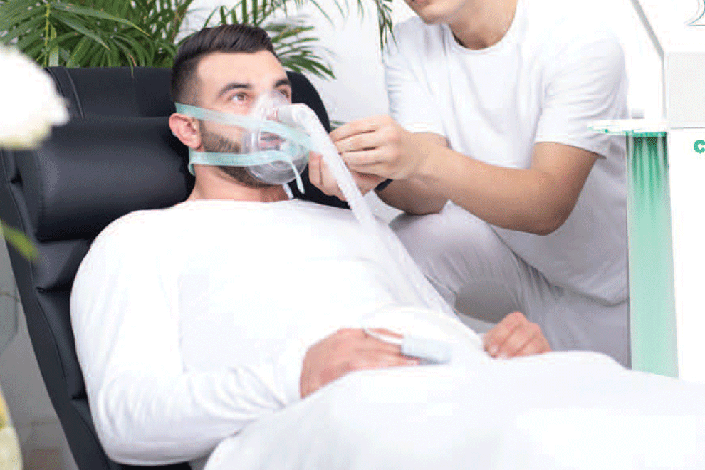 A man lying down wearing an IHHT oxygen mask, with a healthcare worker adjusting it, in a medical setting.