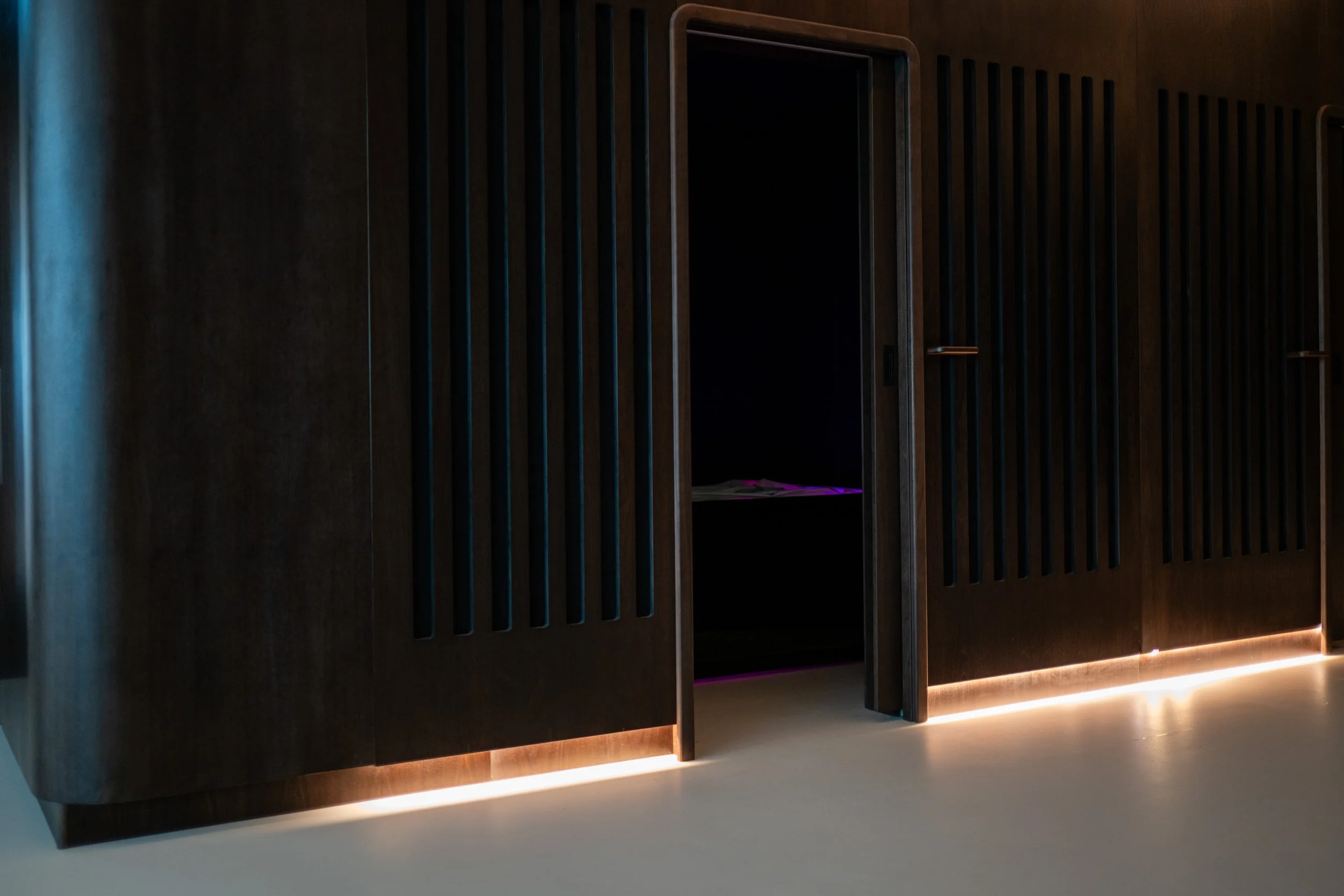 Modern wooden cabinet with vertical slats and a dark interior, illuminated by LED strip lighting at the base, placed on a light-colored floor.