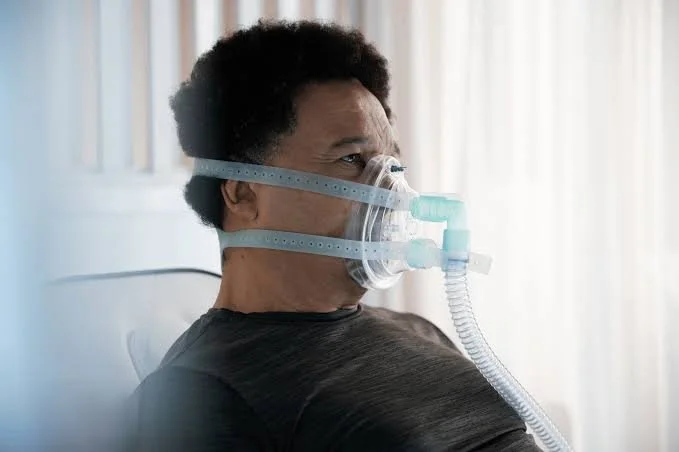 A man wearing a black shirt using a medical oxygen mask connected to a ventilation tube, sitting in a hospital or clinical setting with light curtains in the background.