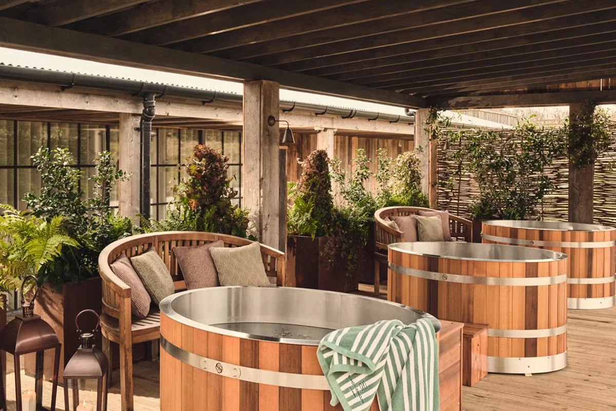 Outdoor kooled ice baths with wooden siding on a wooden deck, surrounded by lush green plants and seating area with pillows, under a dark wooden roof.