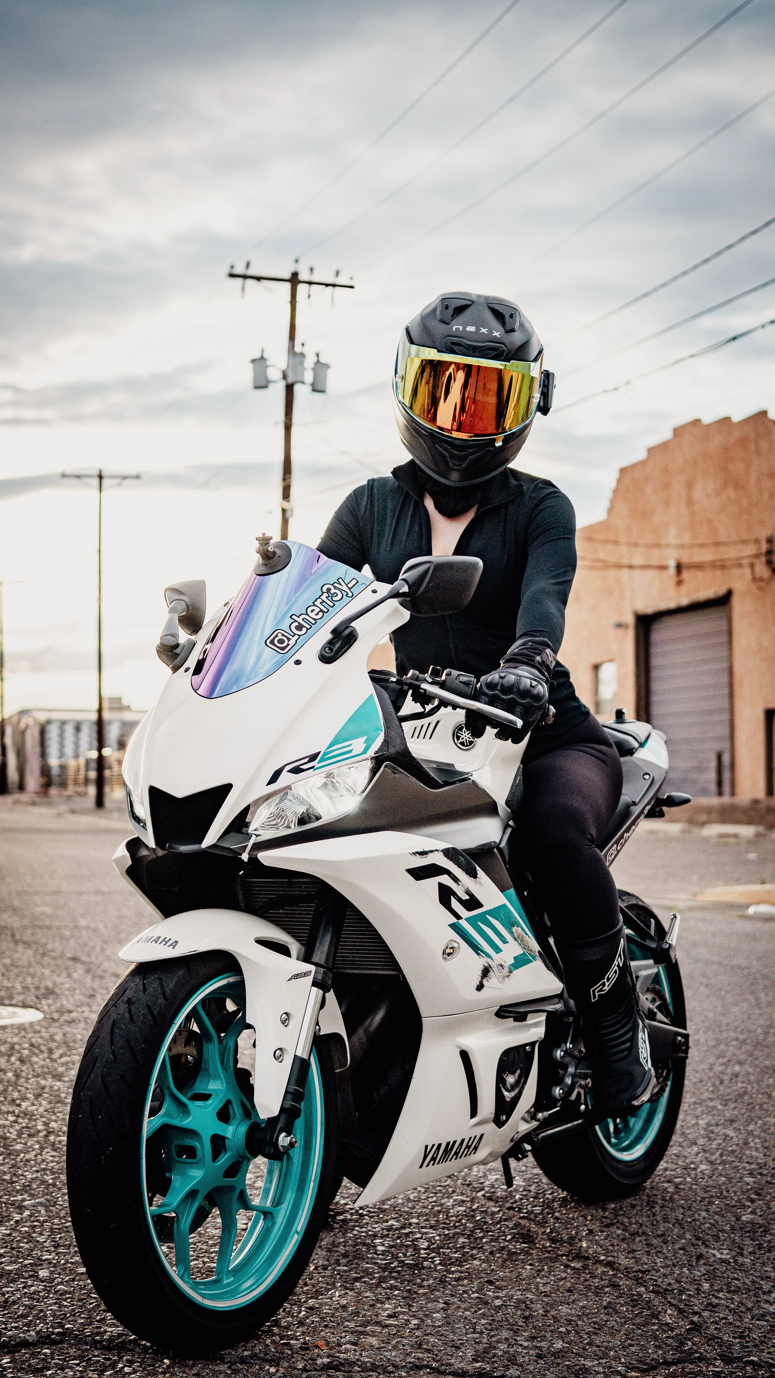 Person wearing a black helmet, black jacket, and gloves sitting on a white Yamaha sport motorcycle with teal accents on a street with a cloudy sky in the background.