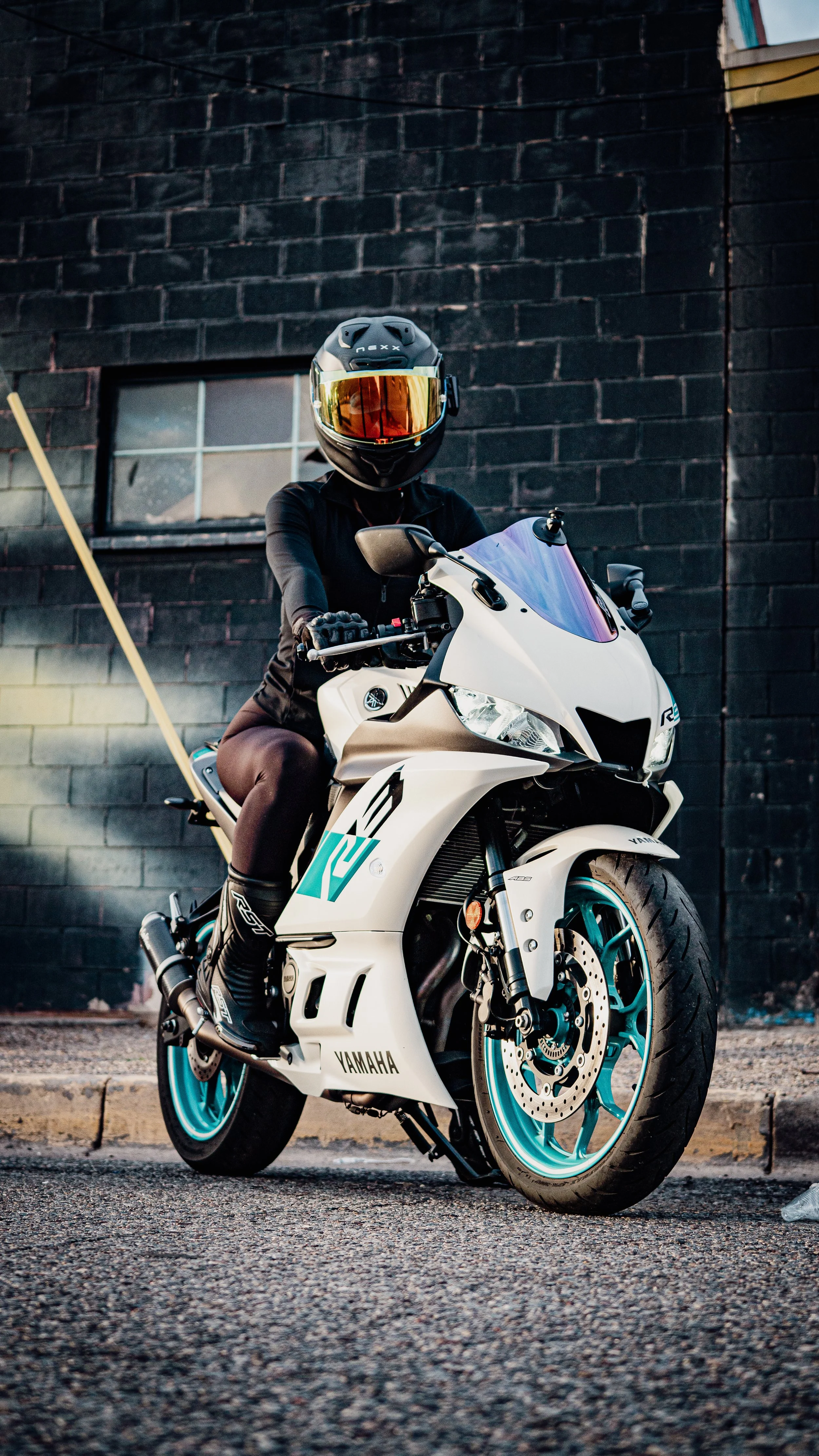 Person wearing black riding gear and helmet, sitting on a white Yamaha sport motorcycle with turquoise accents, parked on a city street with dark brick building and a window in the background.