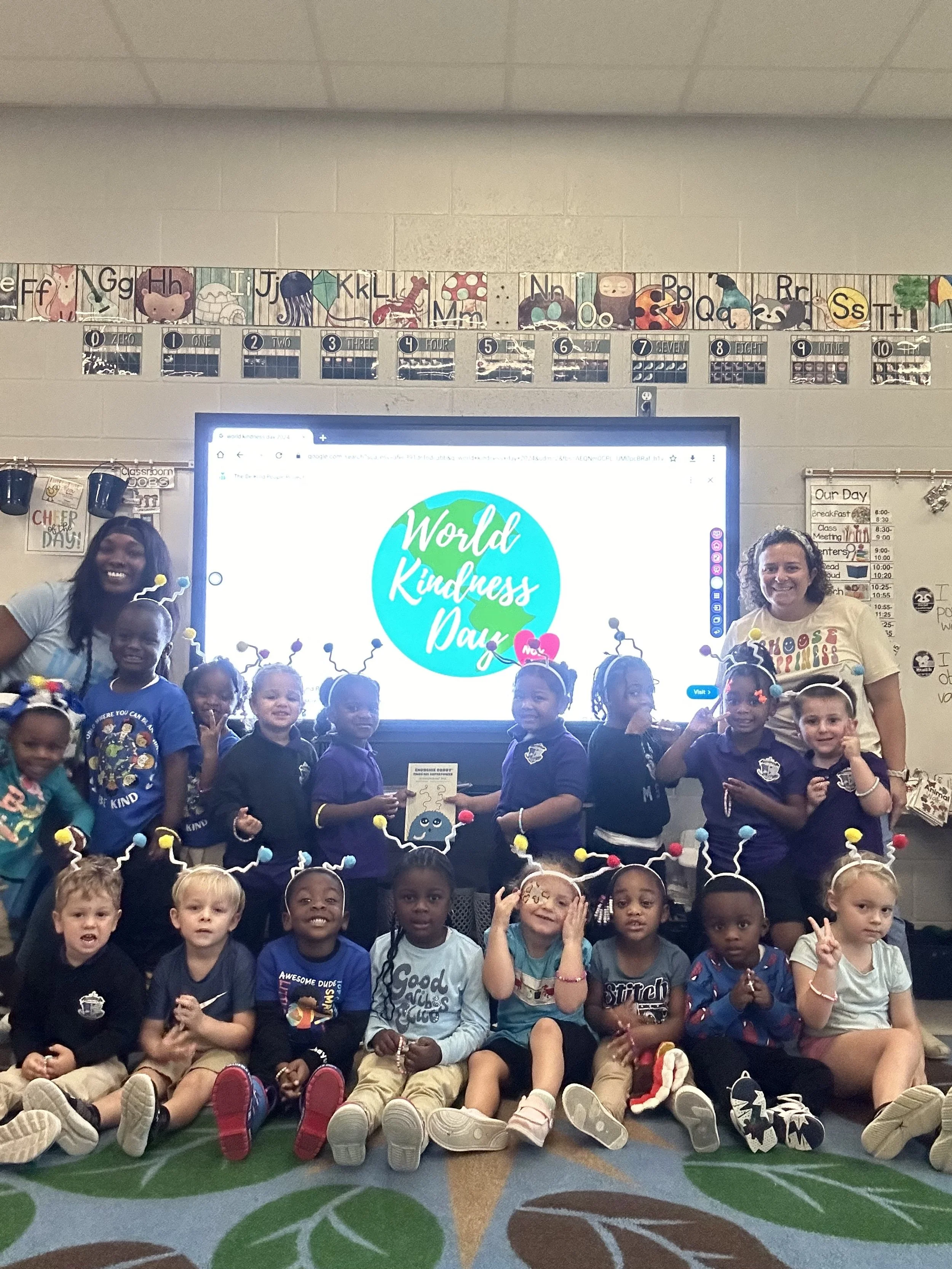 Group of young children and two teachers celebrating World Kindness Day in a classroom, with a large screen displaying the event's logo behind them.