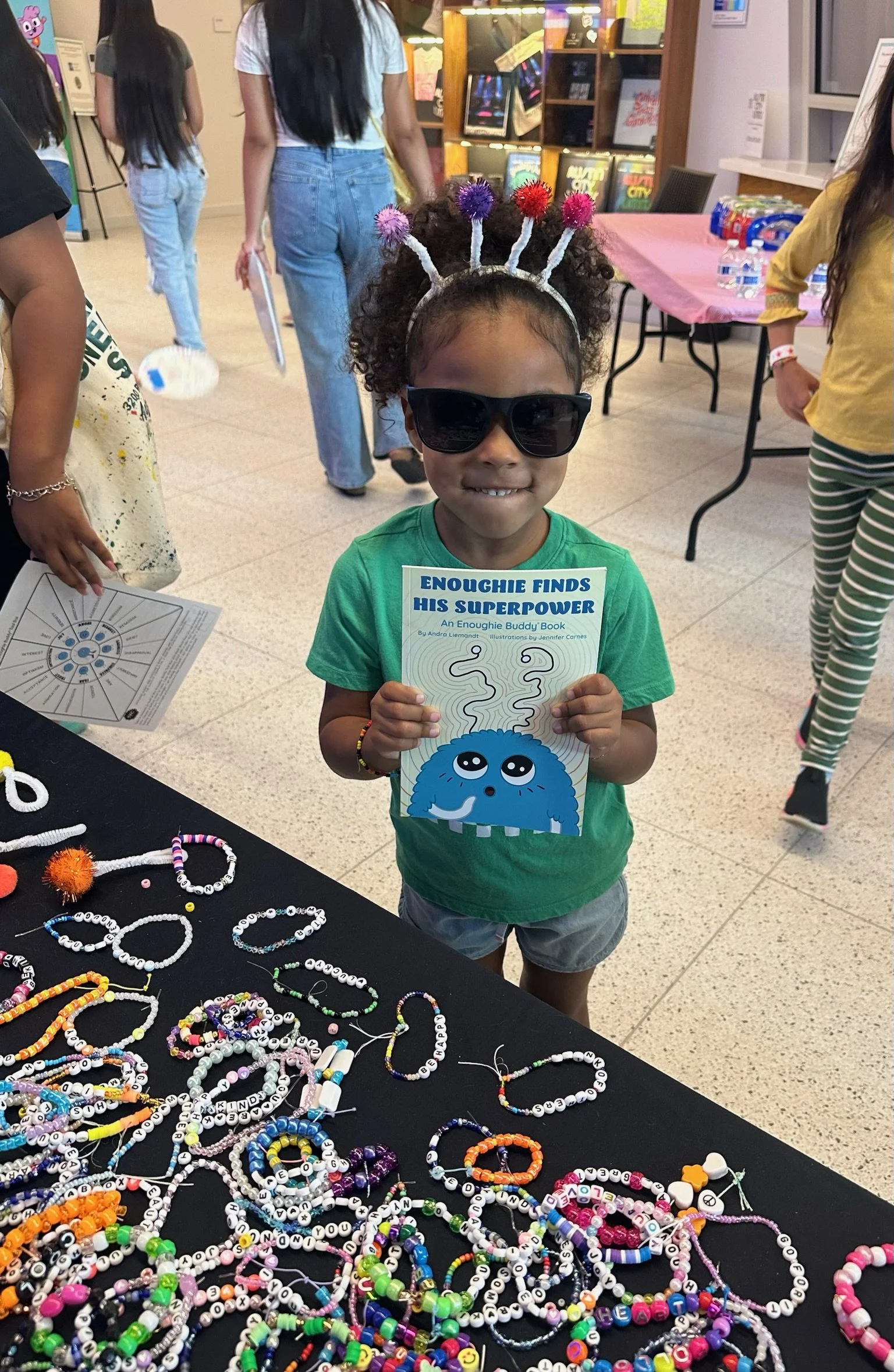 A young girl wearing sunglasses and a headband with fuzzy antennae, smiling and holding a children's book titled 'Enougie Finds His Superpower.' She is standing next to a table covered with multicolored beaded jewelry. Several other children and adul