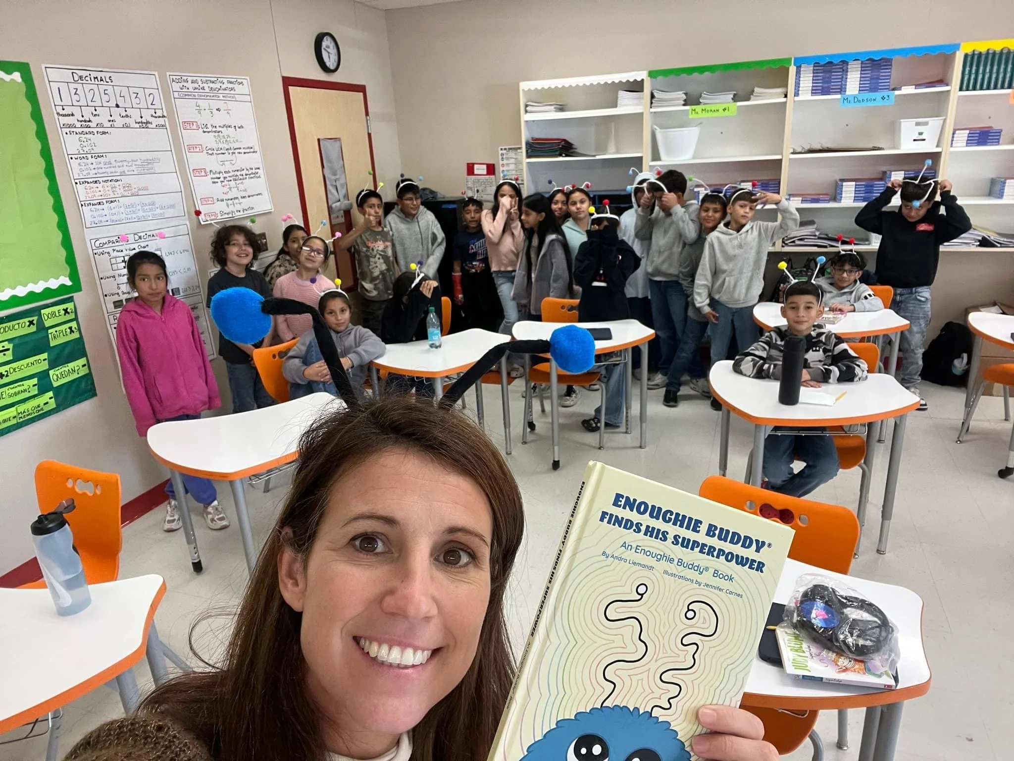 A group of elementary school children in a classroom, some wearing antenna headbands, with a woman in the foreground holding a book titled 'Enoughe Buddy Finds His Superpower' and smiling. Classroom has white shelves, posters, and tables with orange 