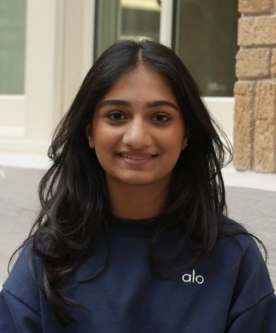 A young woman with long dark hair, smiling, wearing a white sleeveless top and layered gold necklaces, standing against a blue background.