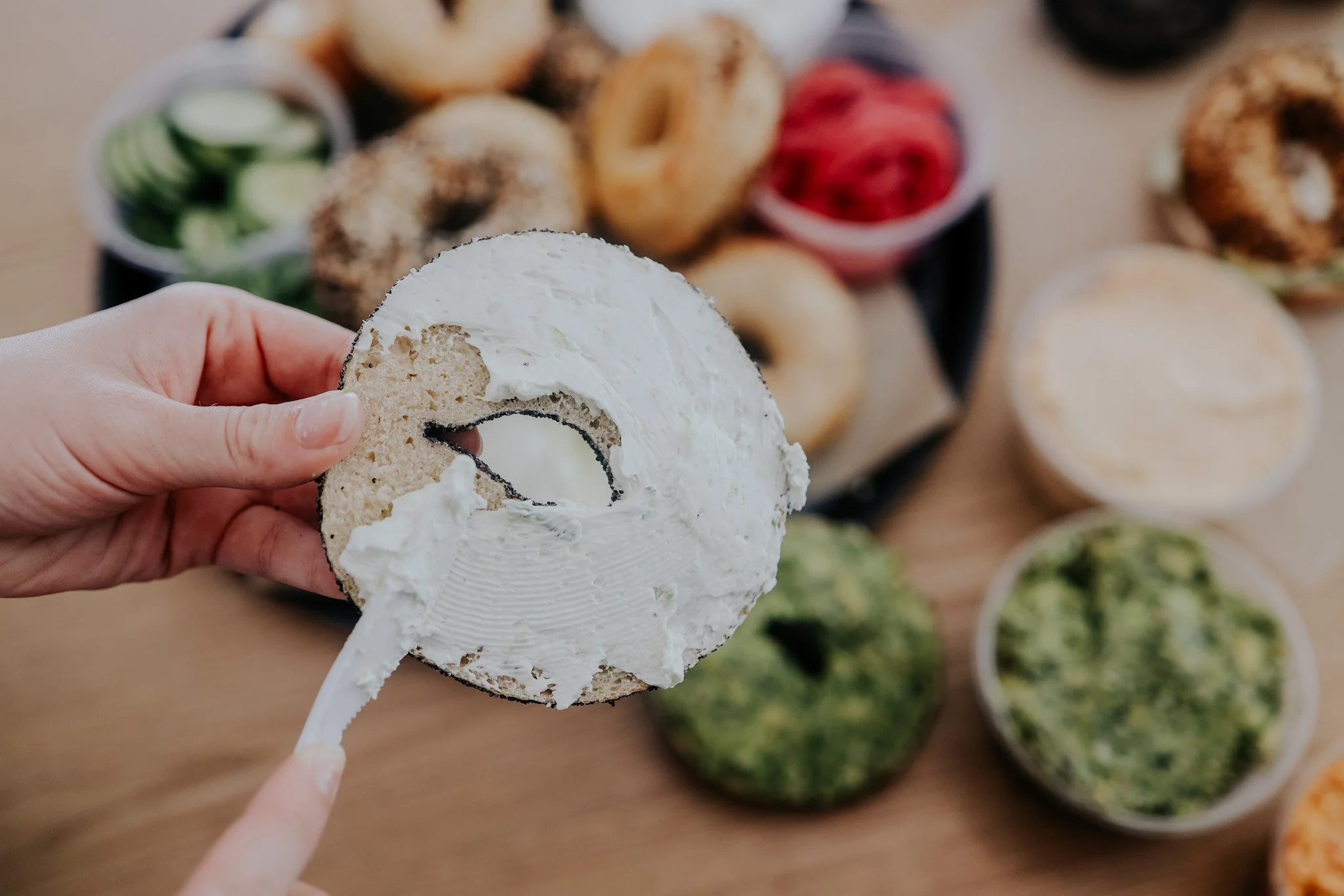 A person spreading cream cheese on a bagel with cucumber slices and other bagels in the background.