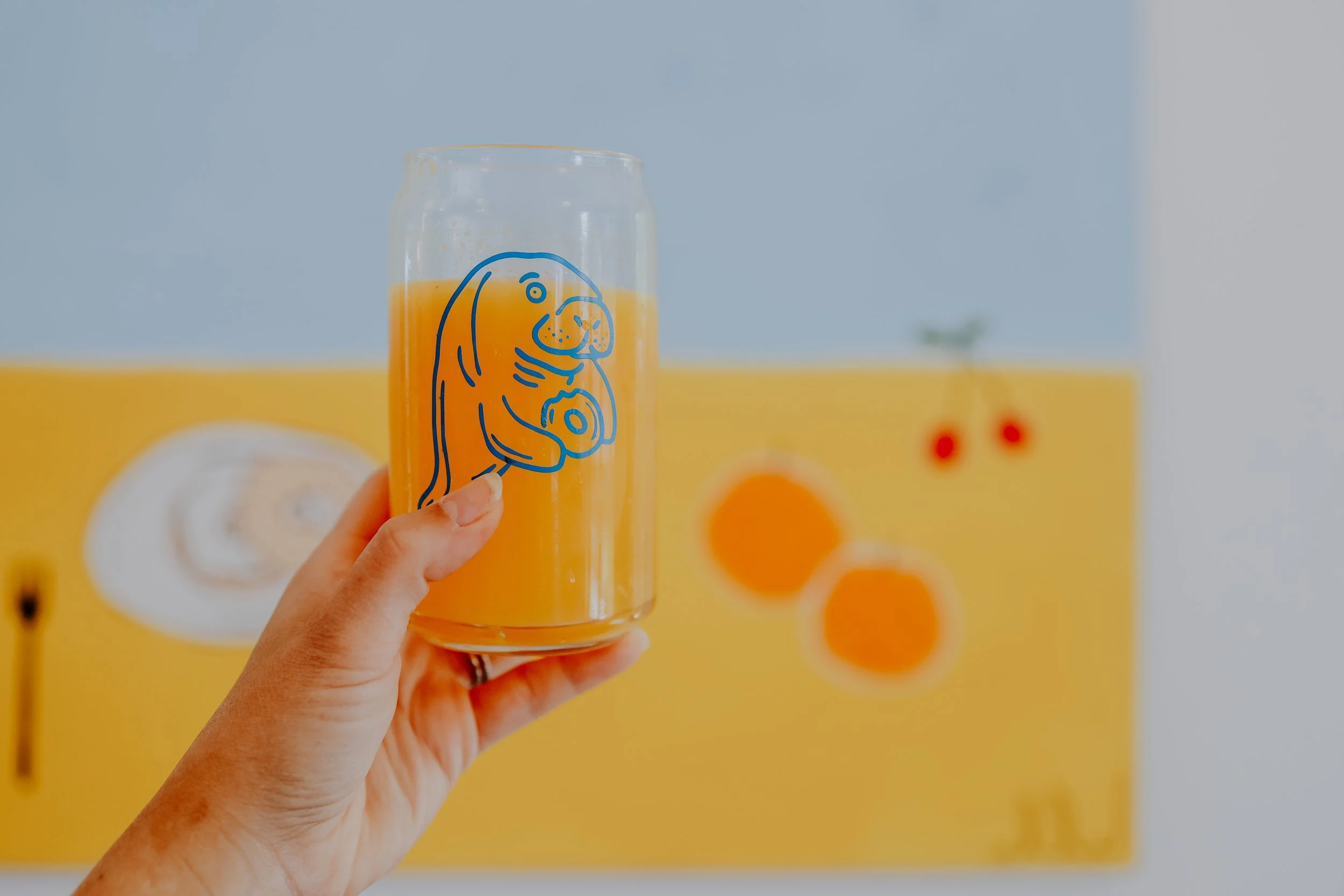 Hand holding a glass filled with orange juice, featuring a blue seal illustration, against a colorful wall background.