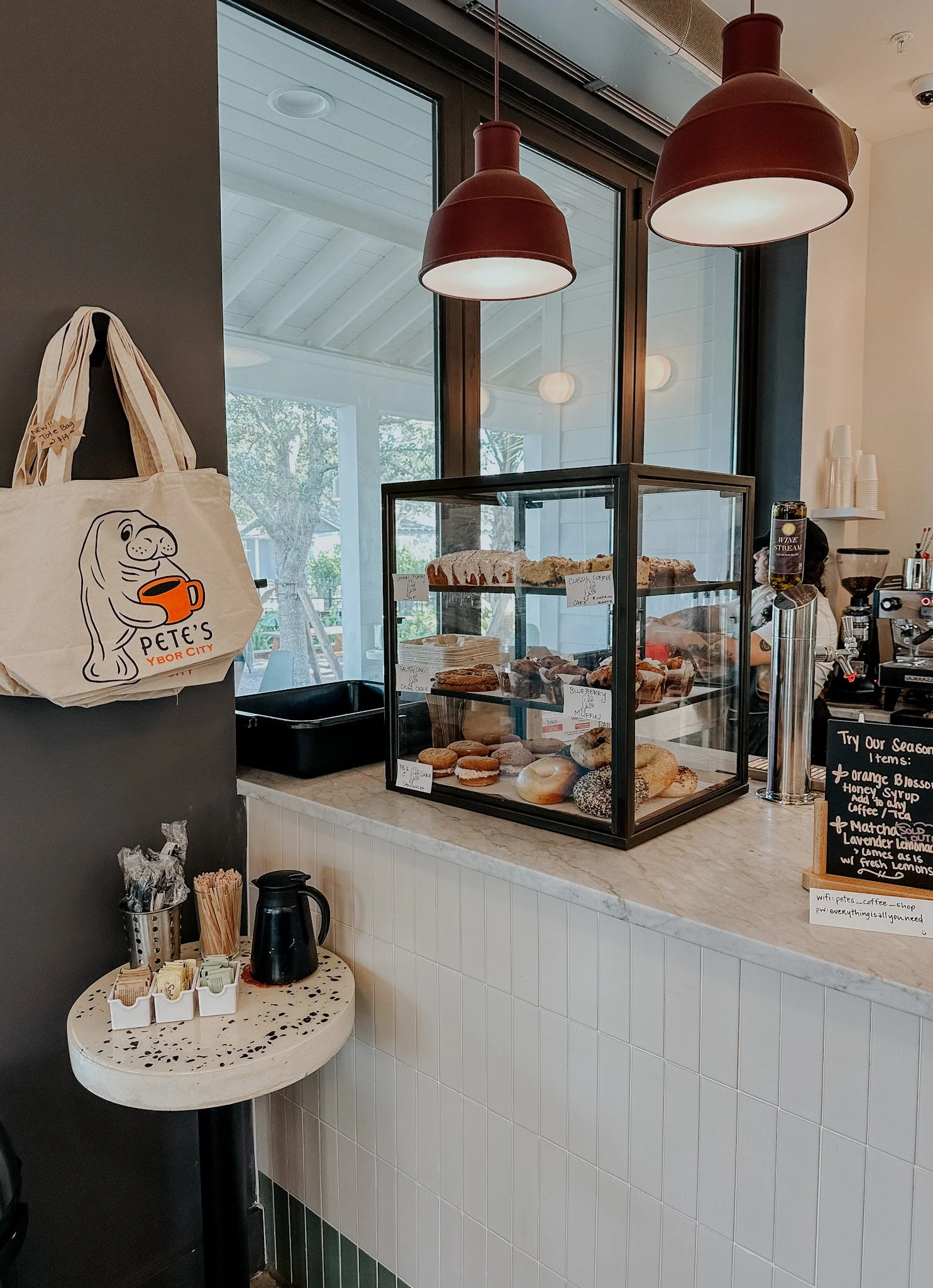 A coffee shop interior featuring a glass display case with assorted pastries and cakes, a hanging tote bag with a seal illustration, red pendant lights, a counter with coffee supplies, and handwritten menu chalkboards.