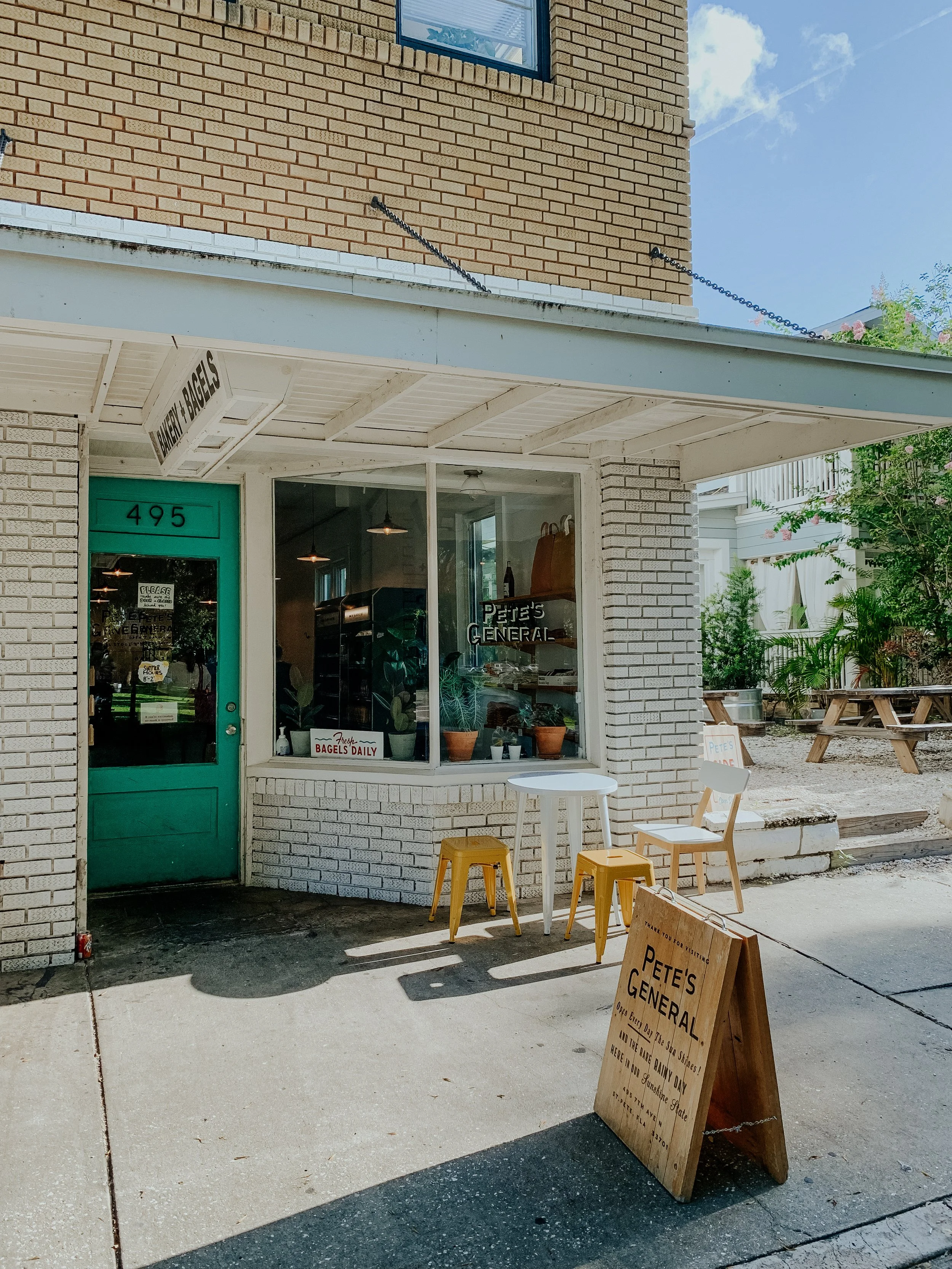 Exterior of a small brick storefront called "Pete's General" with a green door, large glass window, white brick walls, outdoor seating with two yellow chairs and a white table, and a wooden sandwich board sign on the sidewalk.