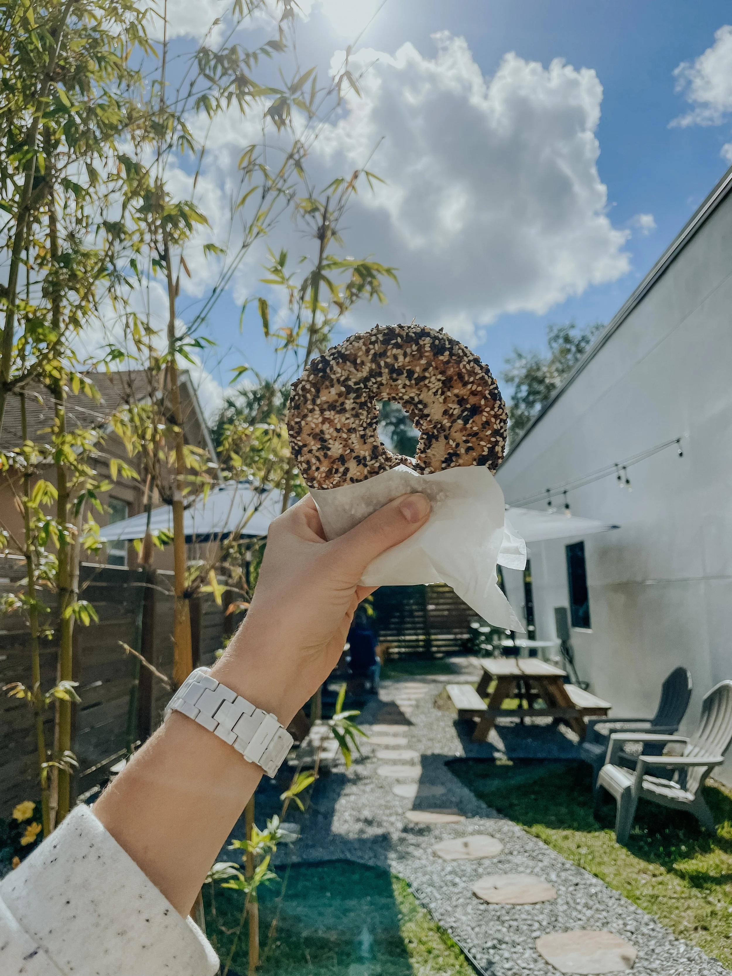 Person holding an everything bagel outside in a sunny garden with a gravel path, picnic table, and scattered patio chairs.