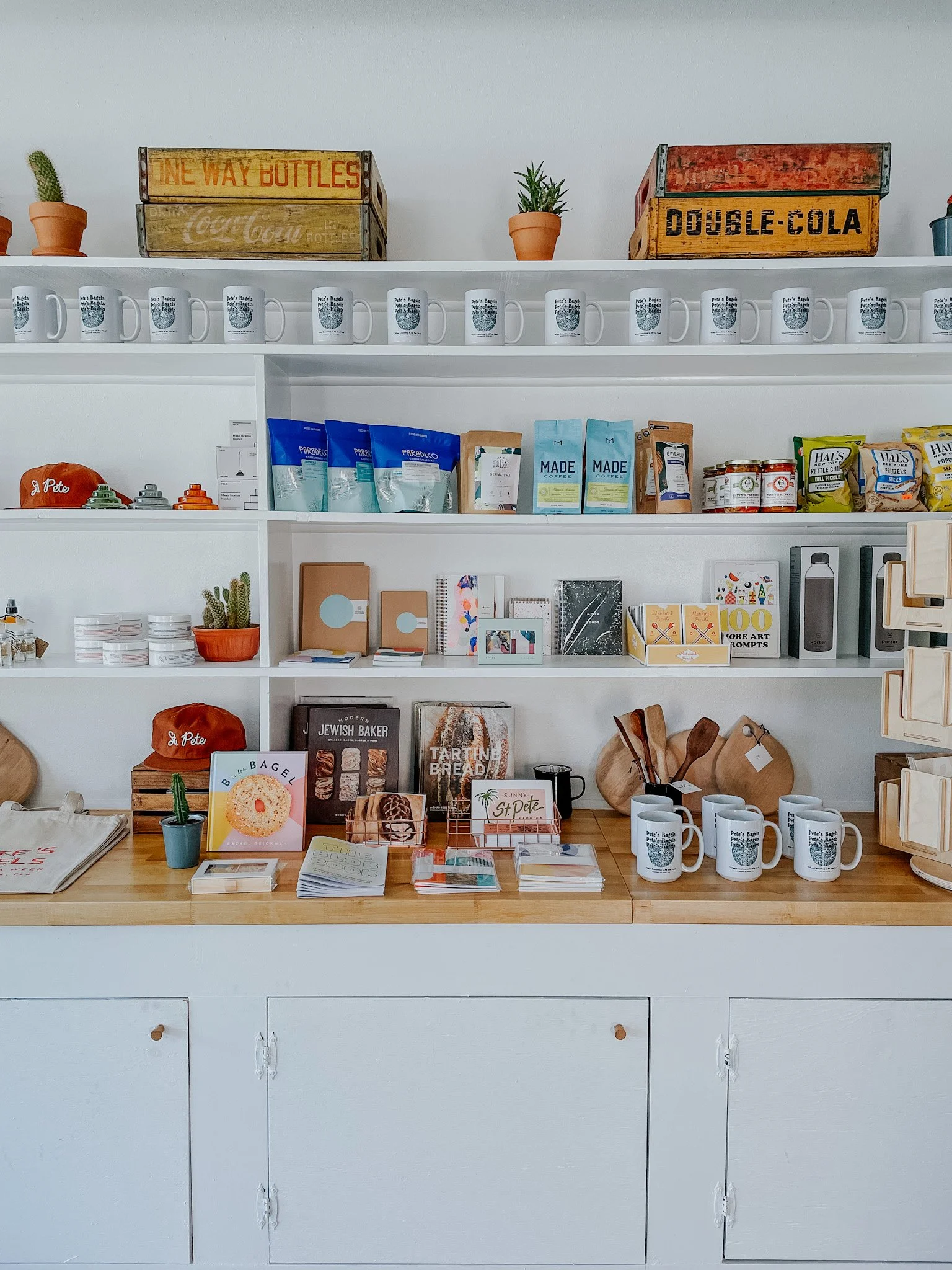 A shop display featuring shelves with various items, including mugs, potted plants, books, snacks, and decorative wooden items. A vintage crate labeled 'One Way Bottles' and 'Double-Cola' is on top. There's also a red cap, notebooks, and kitchen-rela
