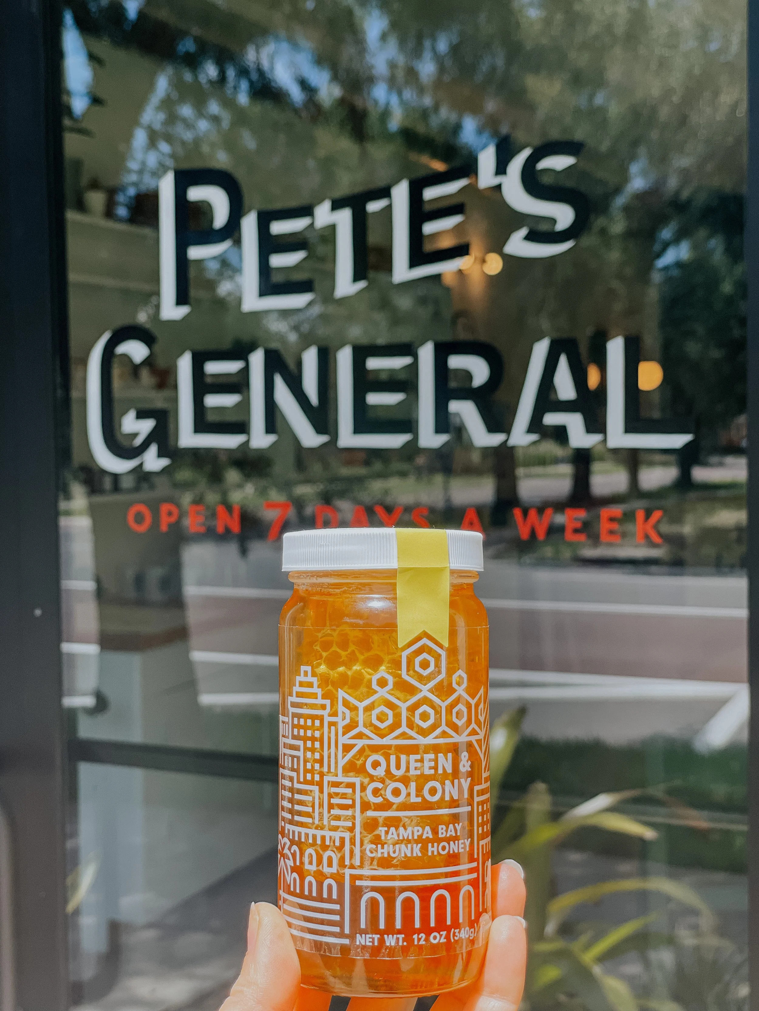 A hand holding a jar of "Queen & Colony Tampa Bay Chunk Honey" in front of a store window that reads "Pete's General Open 7 Days a Week."