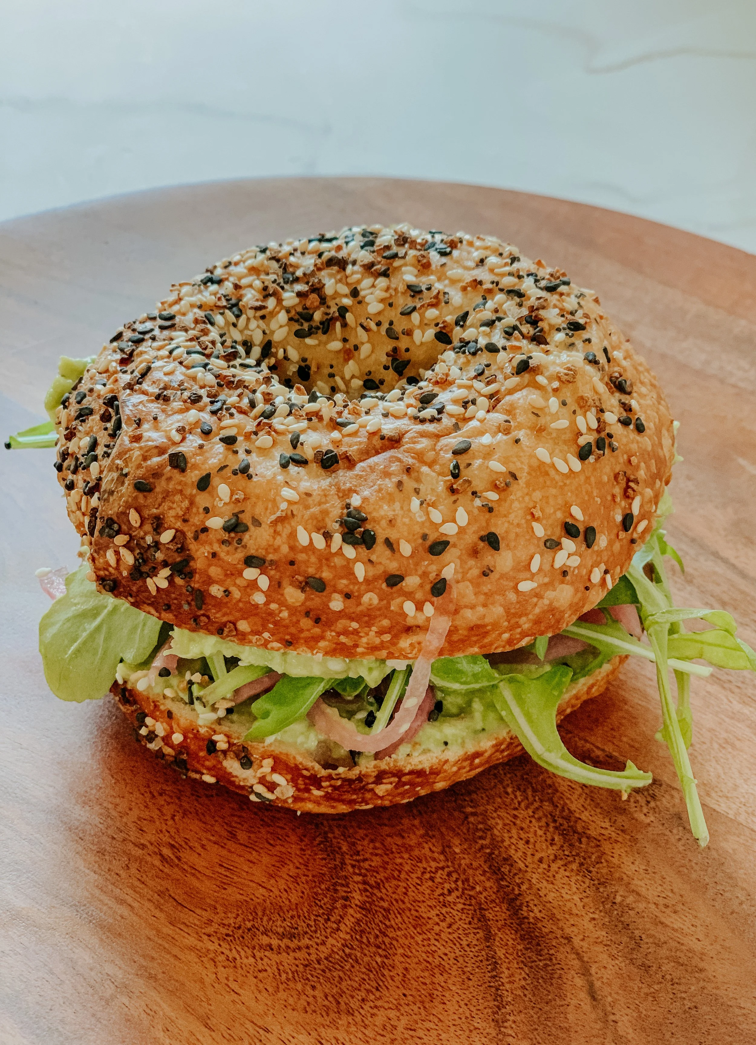 Everything bagel sandwich with greens and avocado on wooden board.