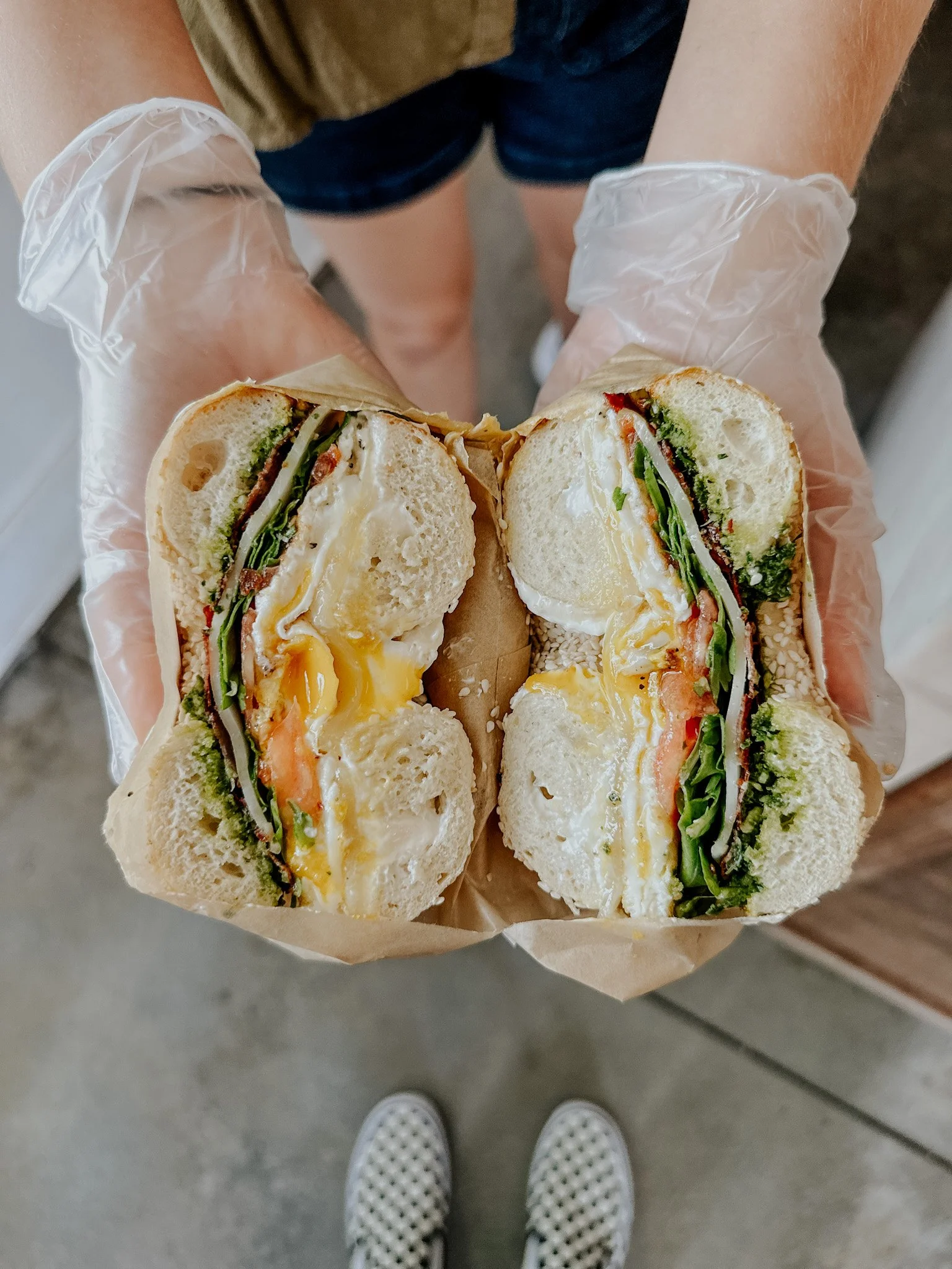 Person holding a halved bagel sandwich with eggs, cheese, tomato, and greens, wearing gloves indoors.