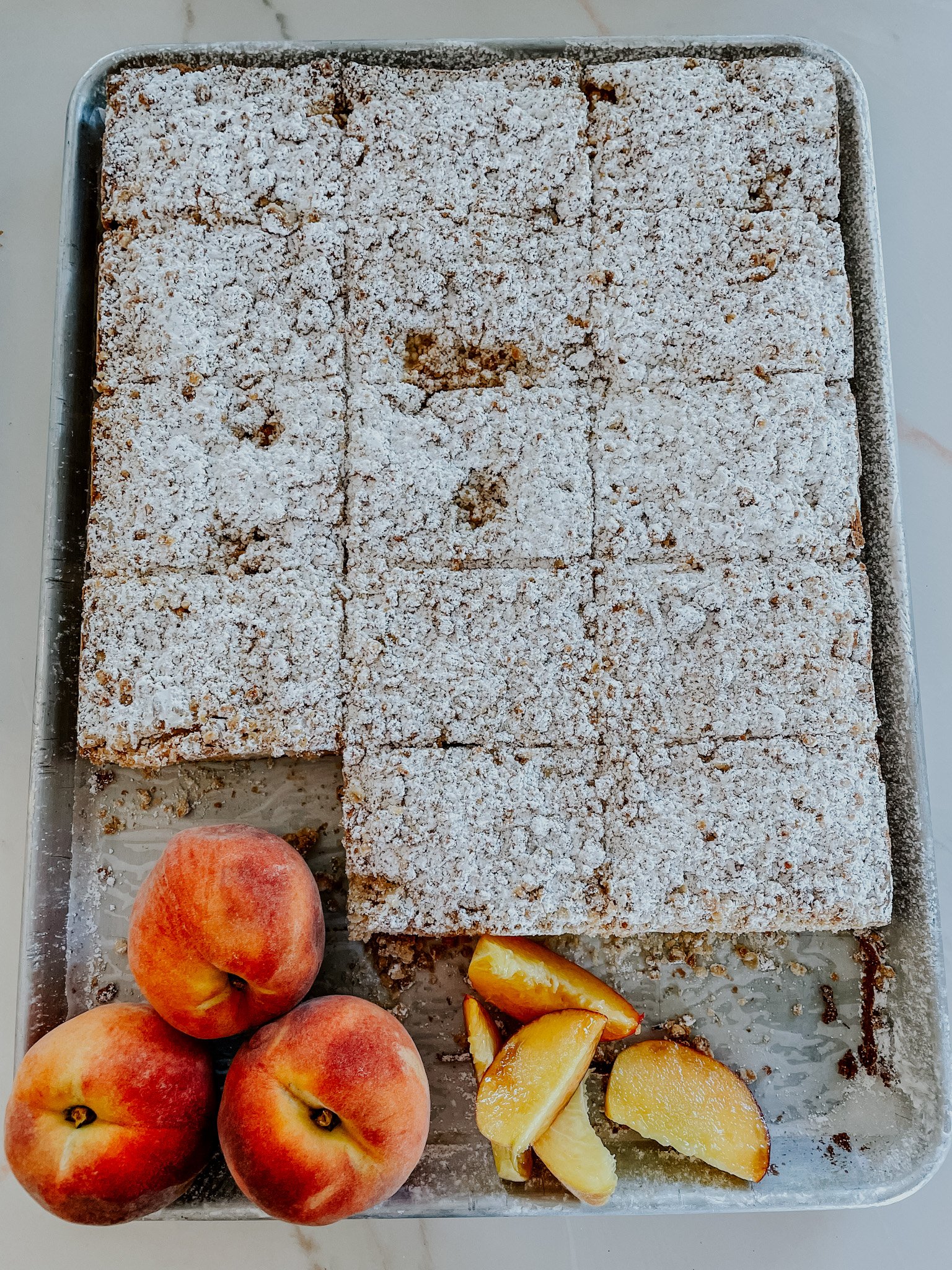 Tray of powdered sugar-topped peach bars with fresh whole and sliced peaches beside them.