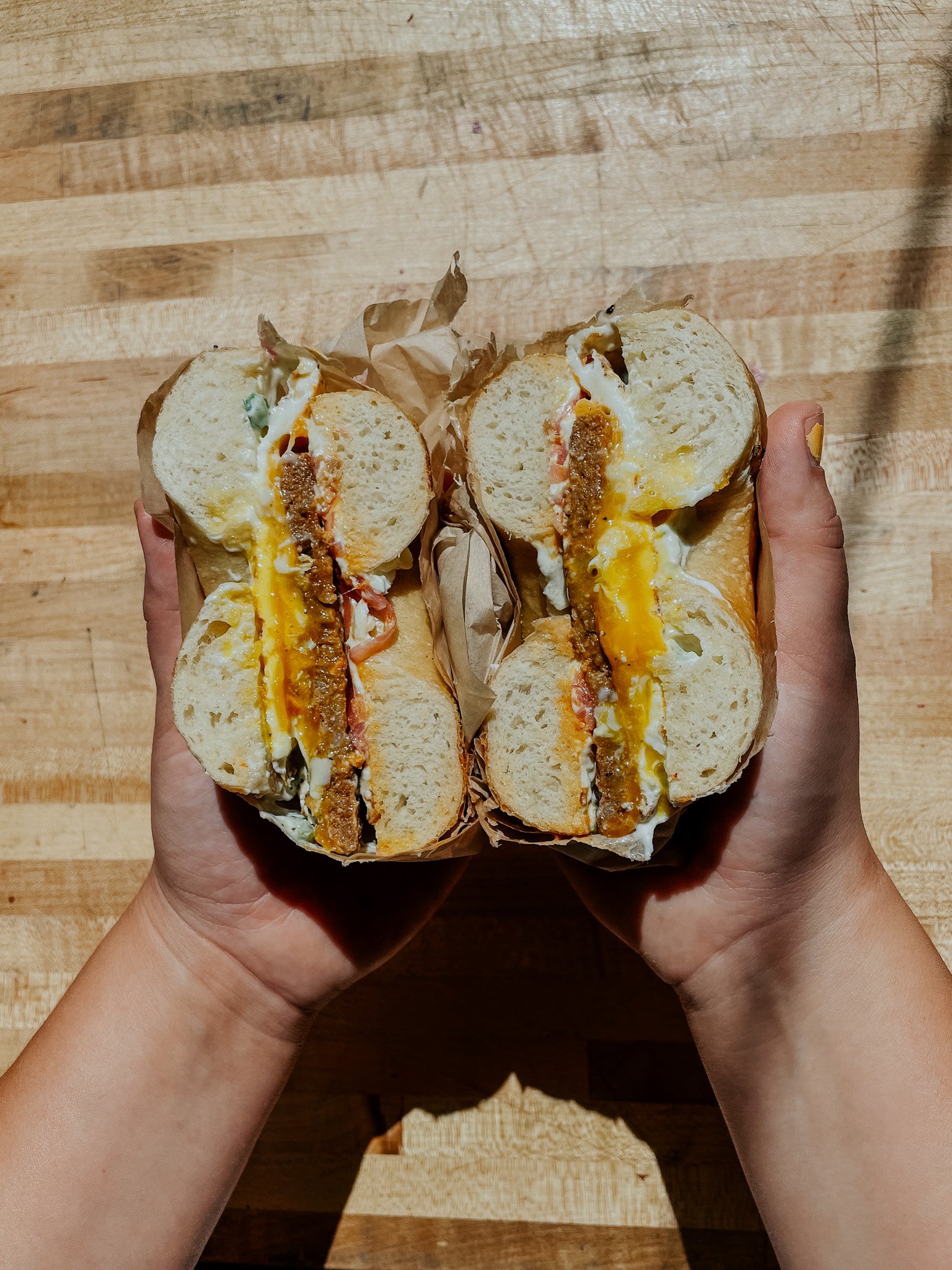 Two halves of a bagel sandwich with egg, cheese, and sausage held by hands on a wooden surface.