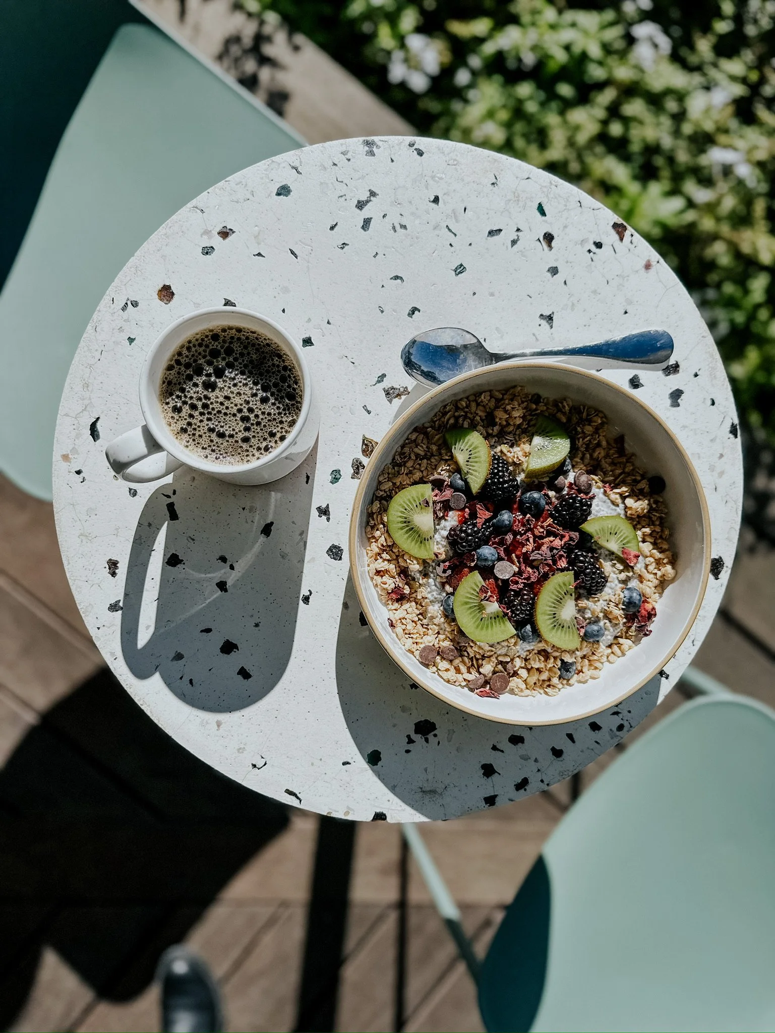 A white circular table with a bowl of granola topped with kiwi slices, blackberries, blueberries, and other fruits, next to a cup of black coffee and a spoon.
