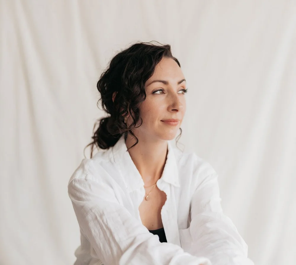 A young woman with dark, curly hair and a nose piercing, wearing a white shirt, looks thoughtfully to the side against a plain, light background.