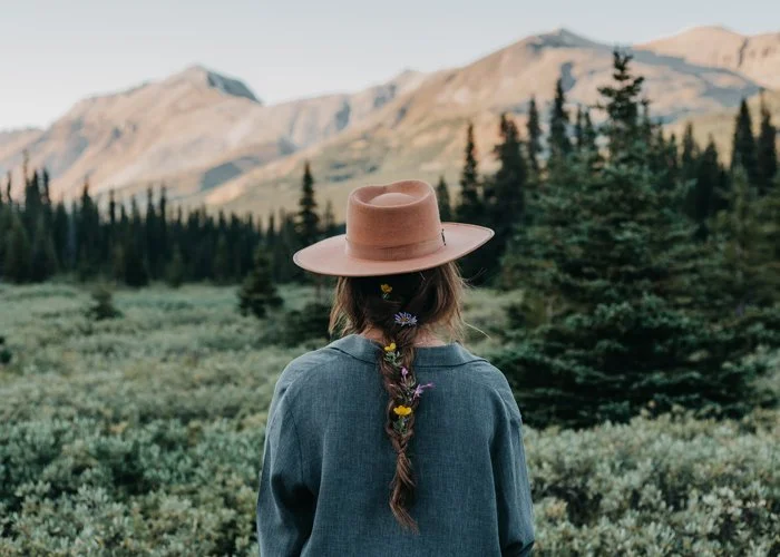 Person wearing a brown hat and denim jacket standing in a green forest with mountains in the background
