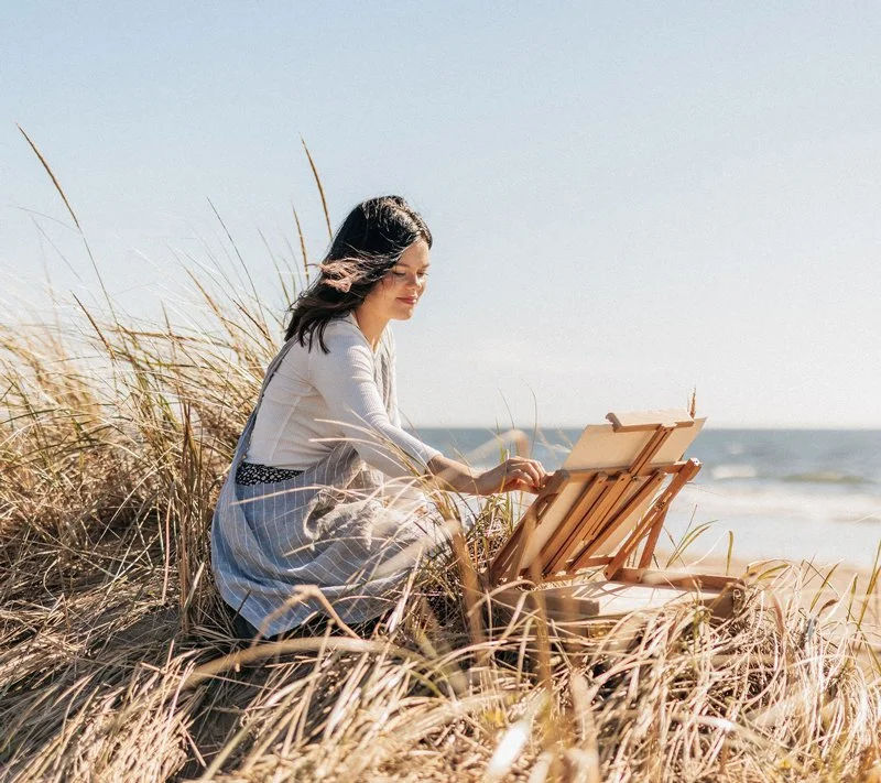 A woman painting on an easel while sitting on a sandy beach surrounded by tall grasses, with the ocean in the background.