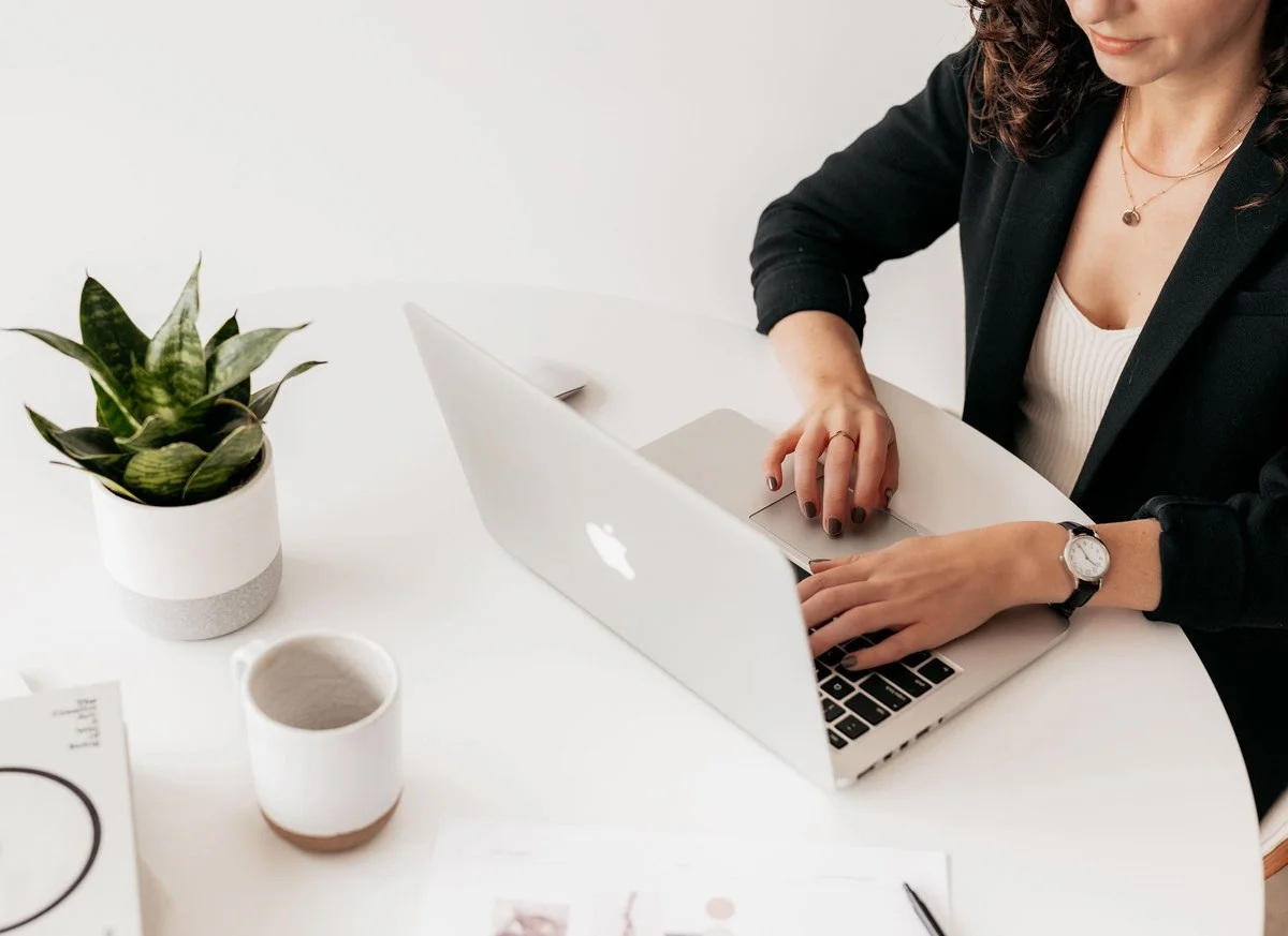 A woman is working on a laptop at a white desk, surrounded by a potted plant, a cup, and some papers.